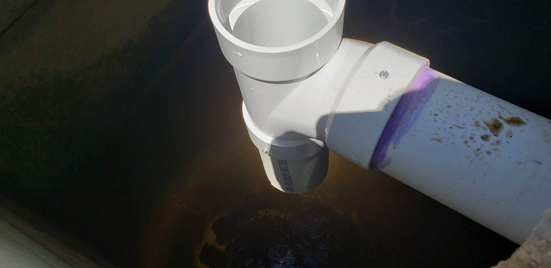 White PVC pipes connected in a dark setting. A gray valve is attached to the bottom pipe.