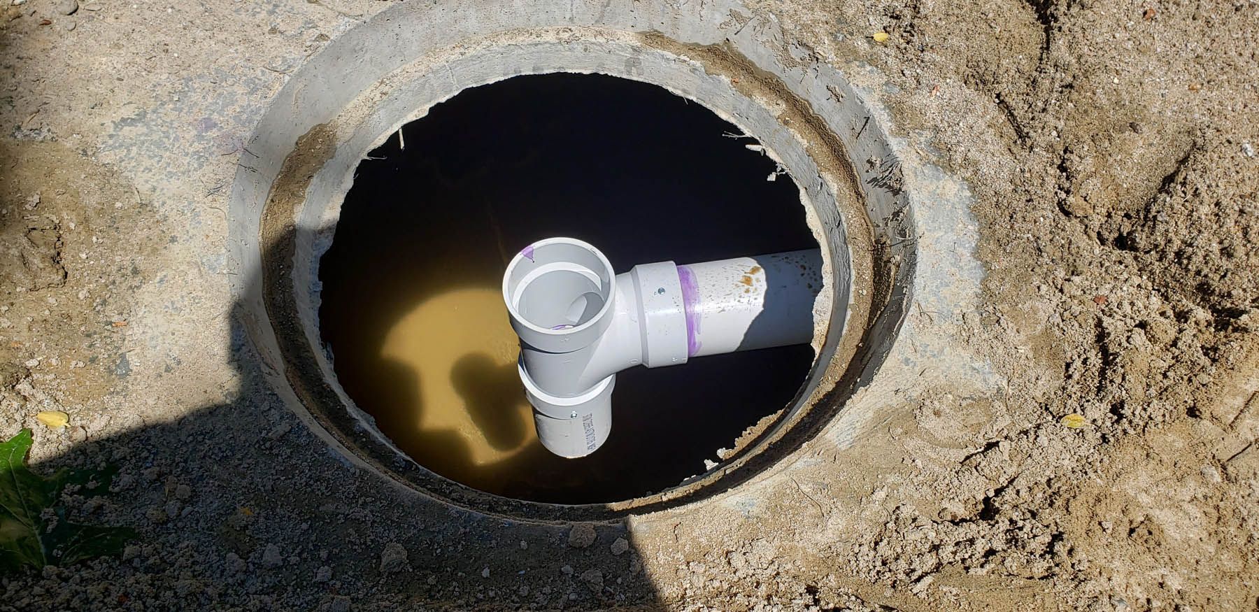 Open manhole with PVC pipe and water inside, surrounded by dirt.