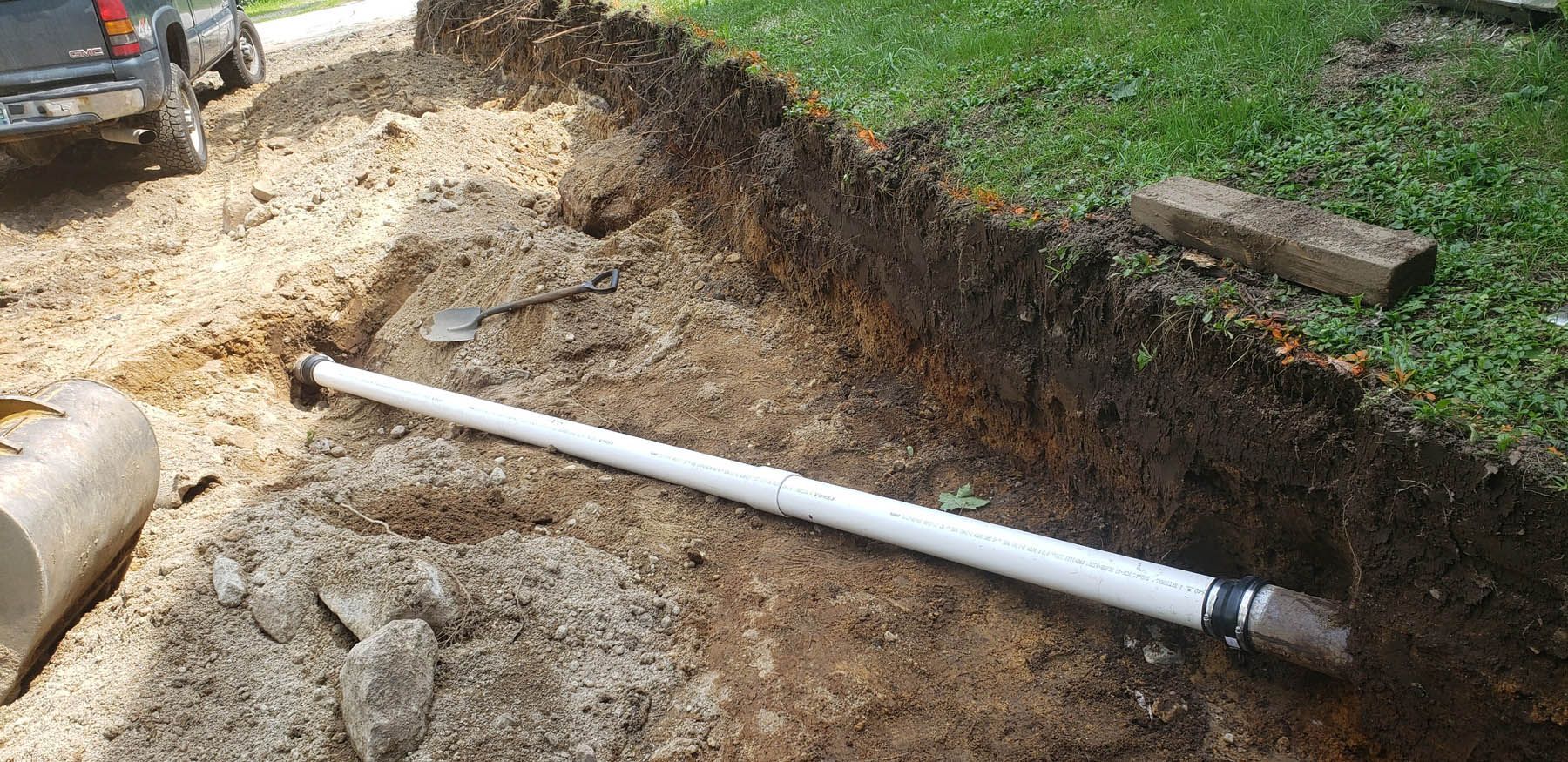 A white PVC pipe is laid in a trench alongside a grass lawn; dirt surrounds.