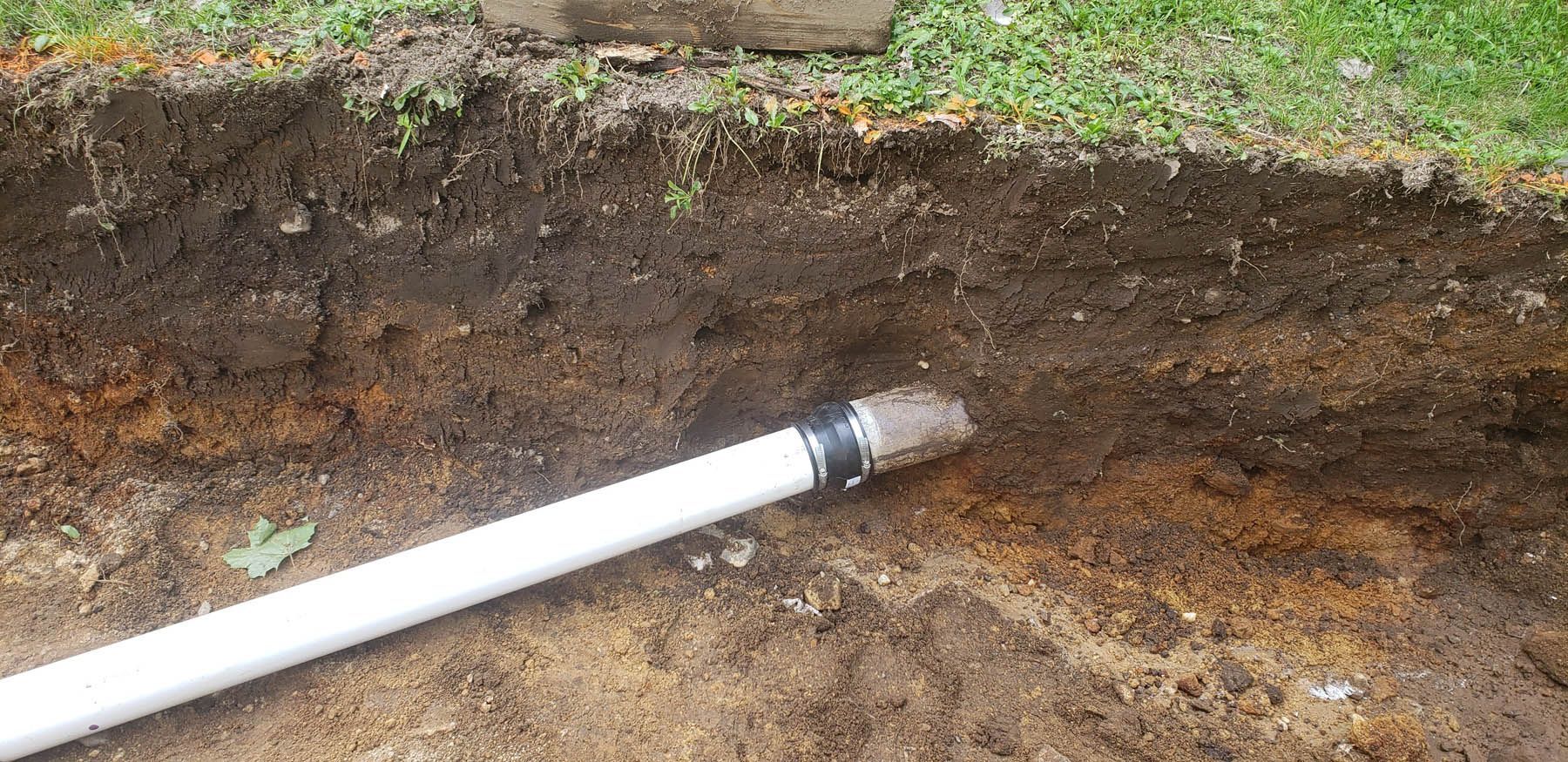 White pipe in a trench, ready to be buried. Dirt, grass, and soil are visible.
