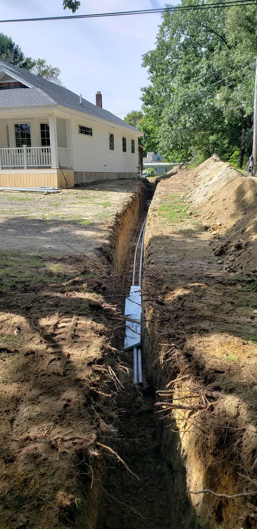 A trench dug in the ground with white pipes and electrical box, beside a house on a sunny day.