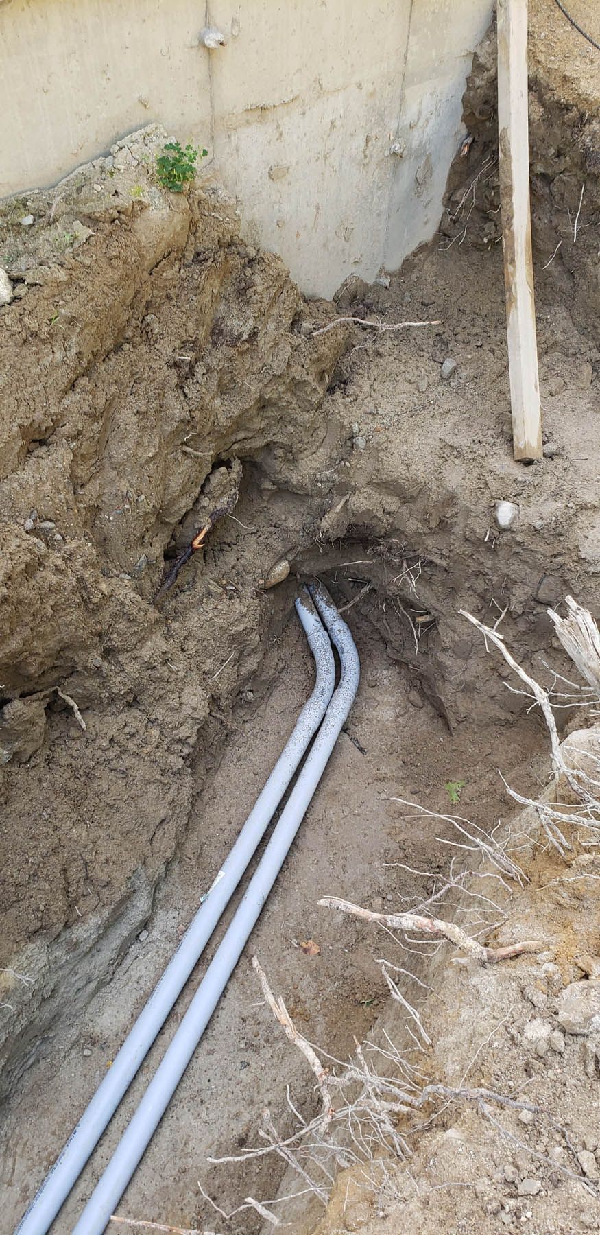Two gray pipes in a dirt trench near a concrete wall, likely for utilities.