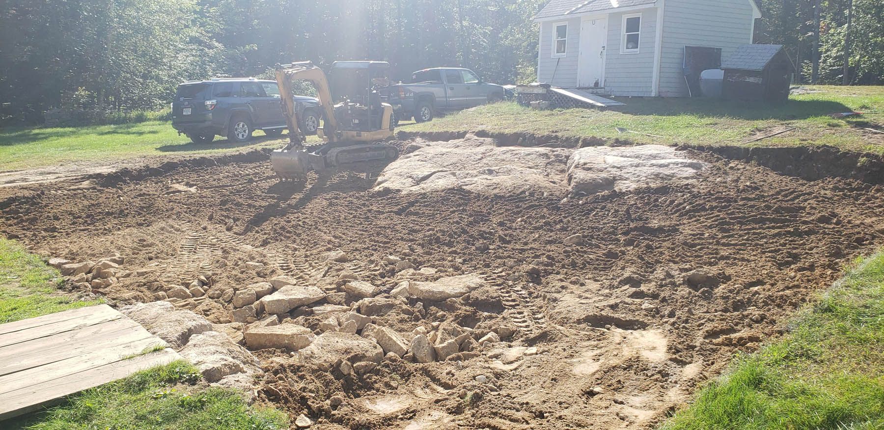 A large hole dug in a yard with an excavator, a house, and vehicles in the background.