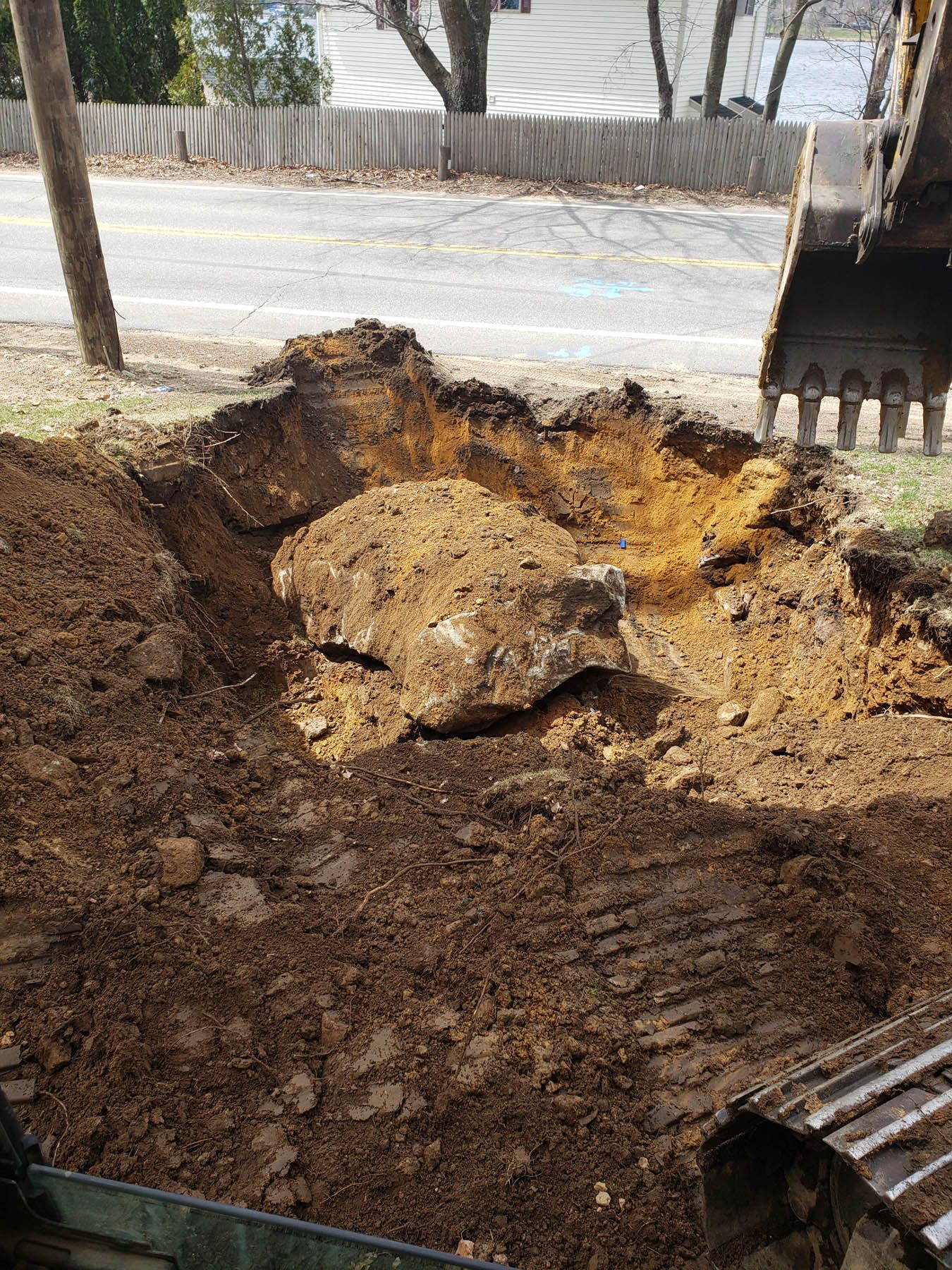 An excavator digging in the ground near a road; a large rock is exposed in the hole.