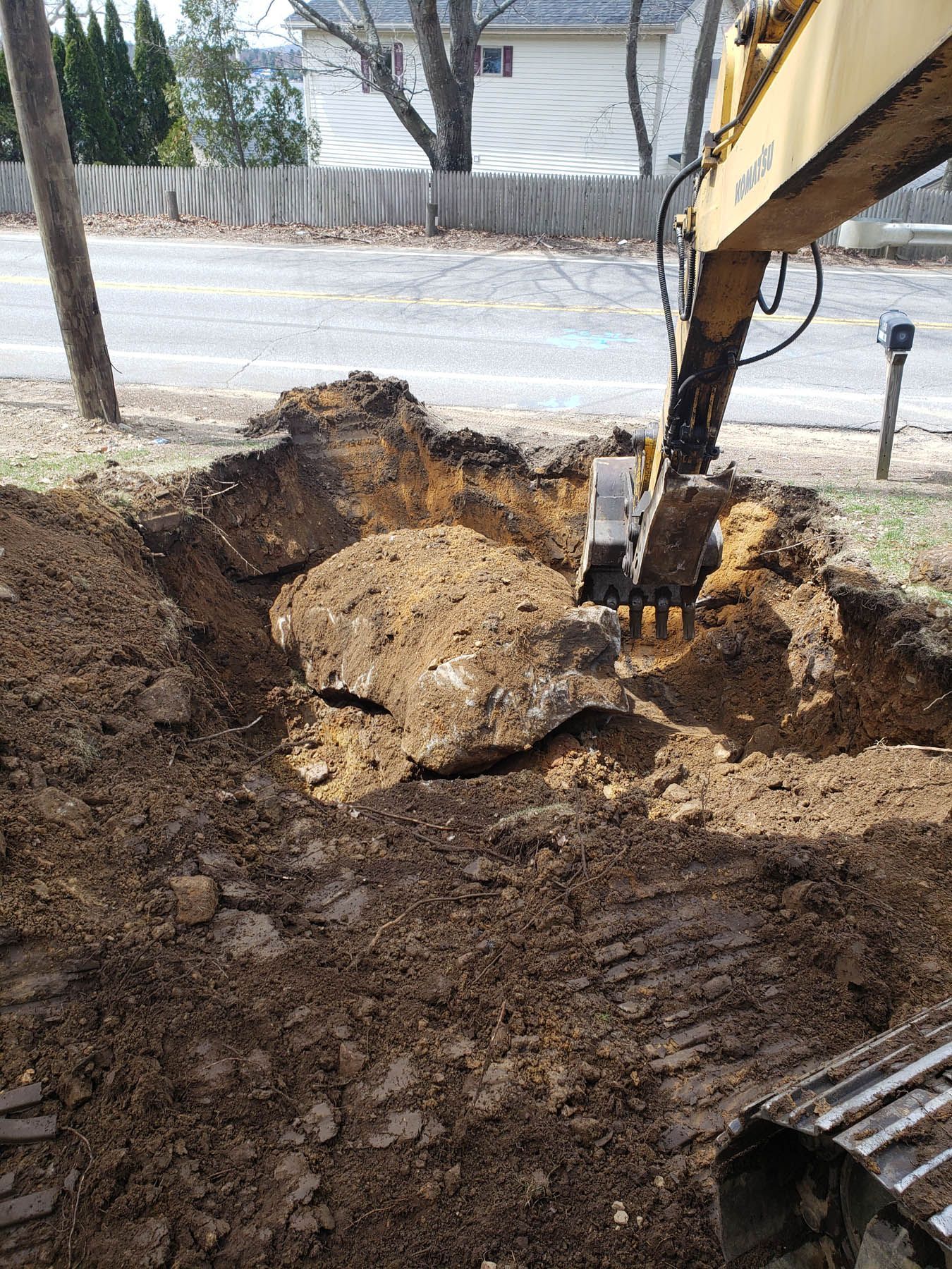 An excavator digs a large hole in a yard, revealing soil and asphalt.