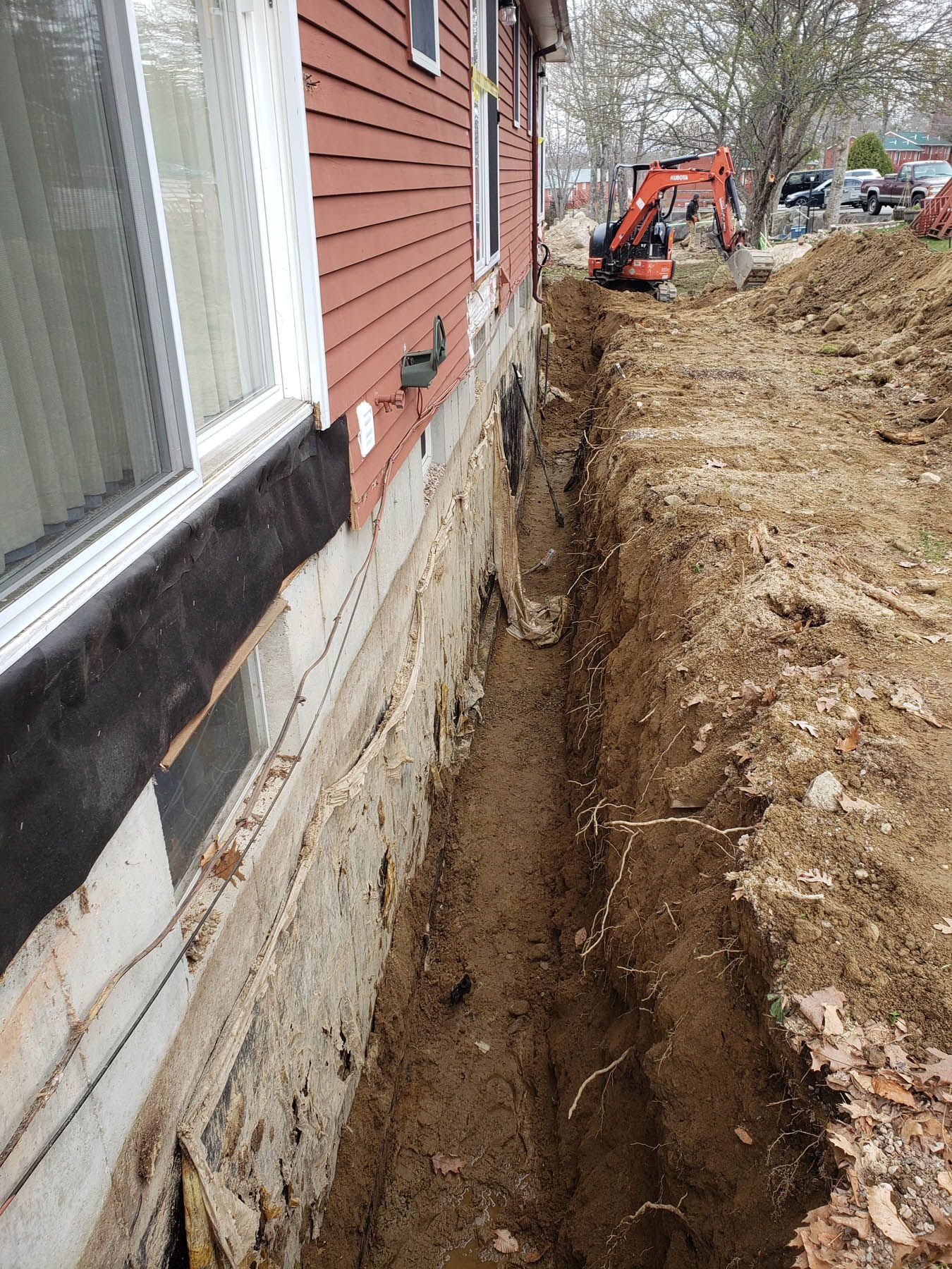 Trench dug along a red-sided building's foundation. An excavator is visible in the distance.