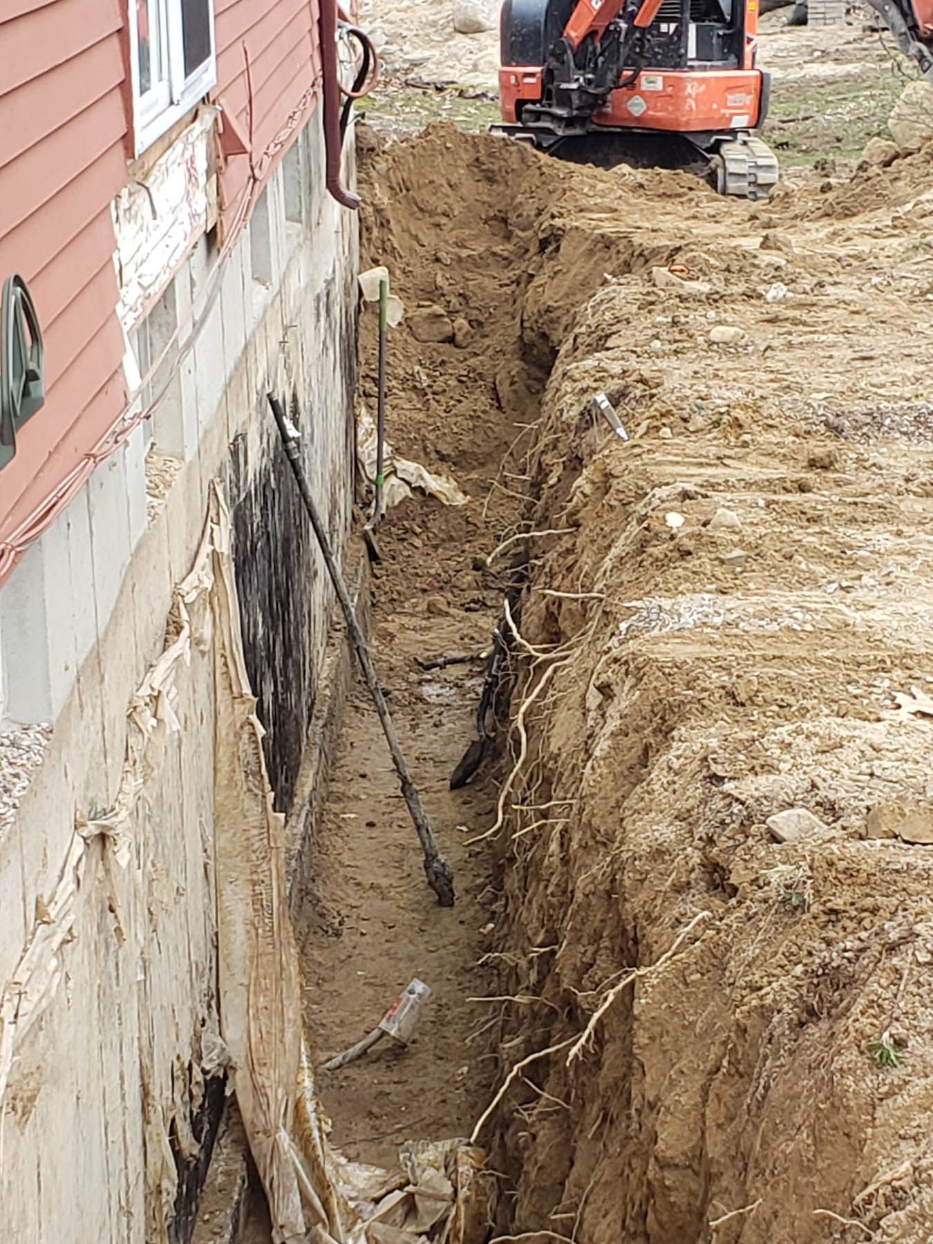 Trench dug next to a house foundation. An orange excavator is in the background.