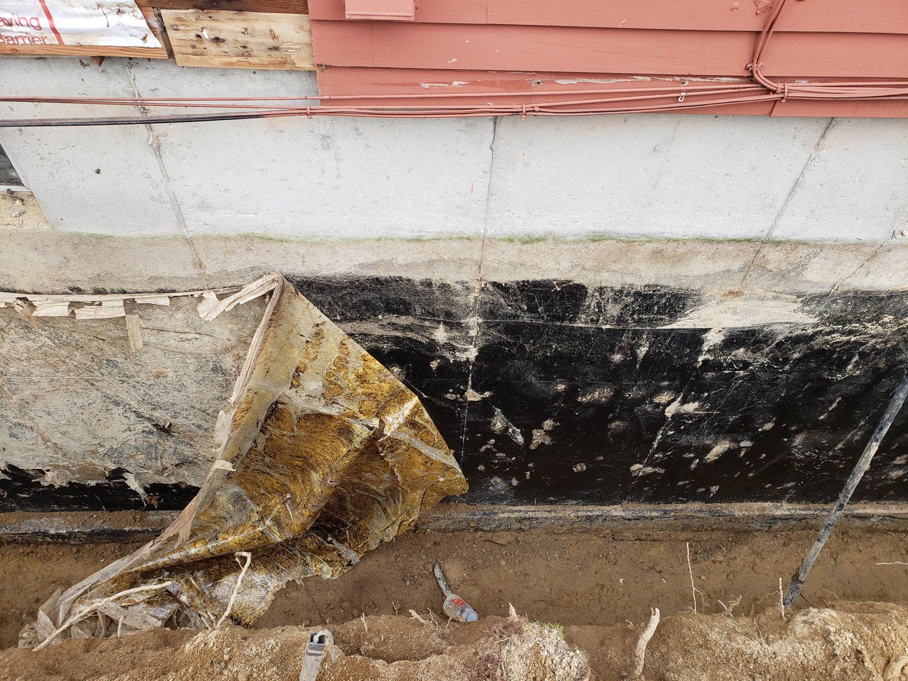 Foundation of a building with exposed black sealant and a torn, brown tarp near the ground.