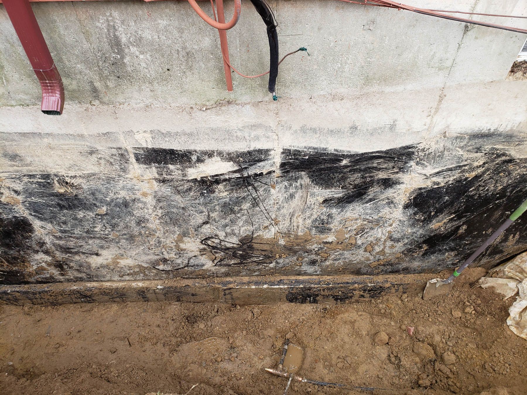 Foundation with black waterproofing, dirt, and exposed pipes.