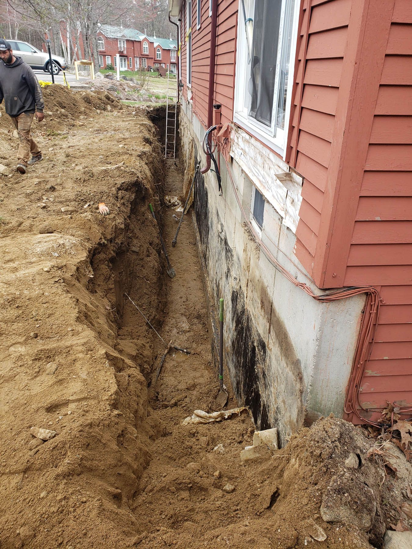 A trench dug next to a red house. A person walks nearby.