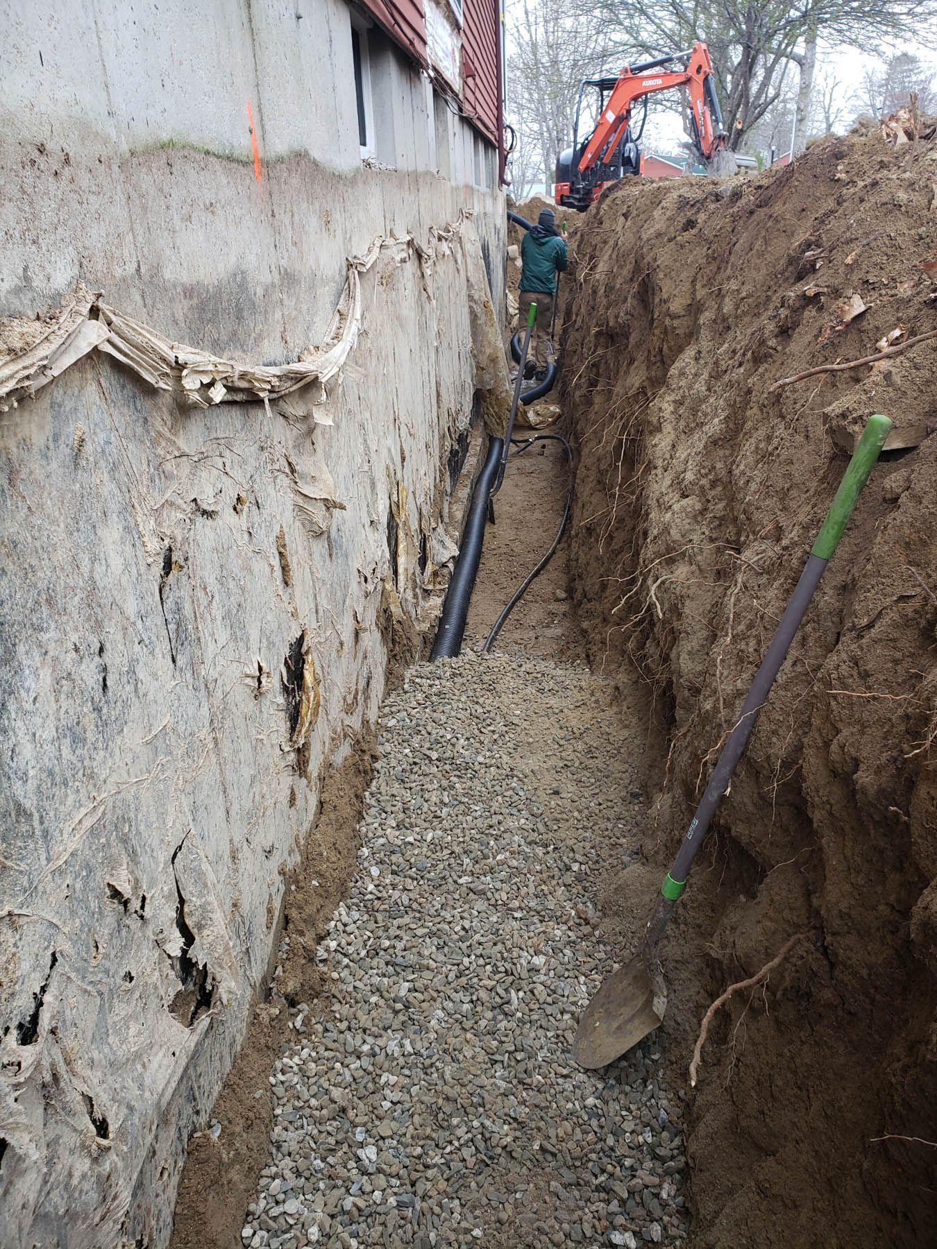 A narrow trench with gravel, pipes, and a foundation wall. An excavator and worker are visible in the background.