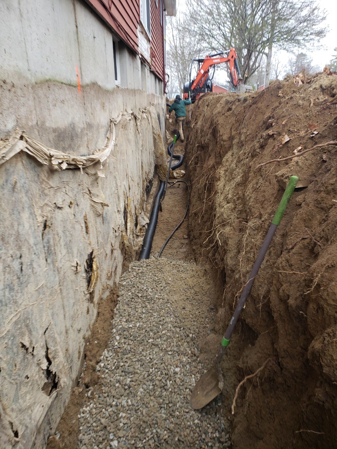 Trench beside a building with gravel, pipes, and a shovel. An excavator works in the background.