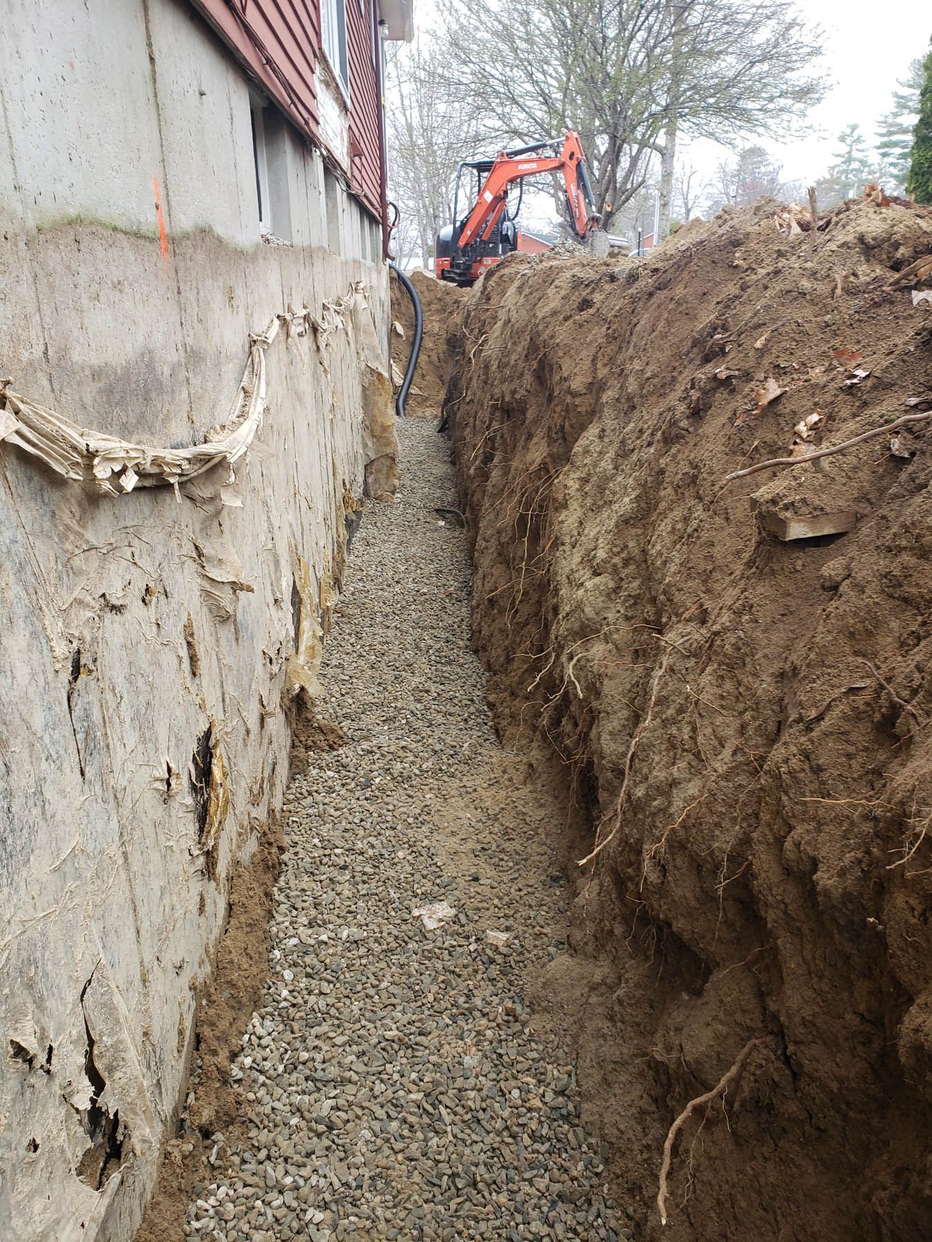 Gravel-lined trench next to a building foundation, with an excavator in the background.