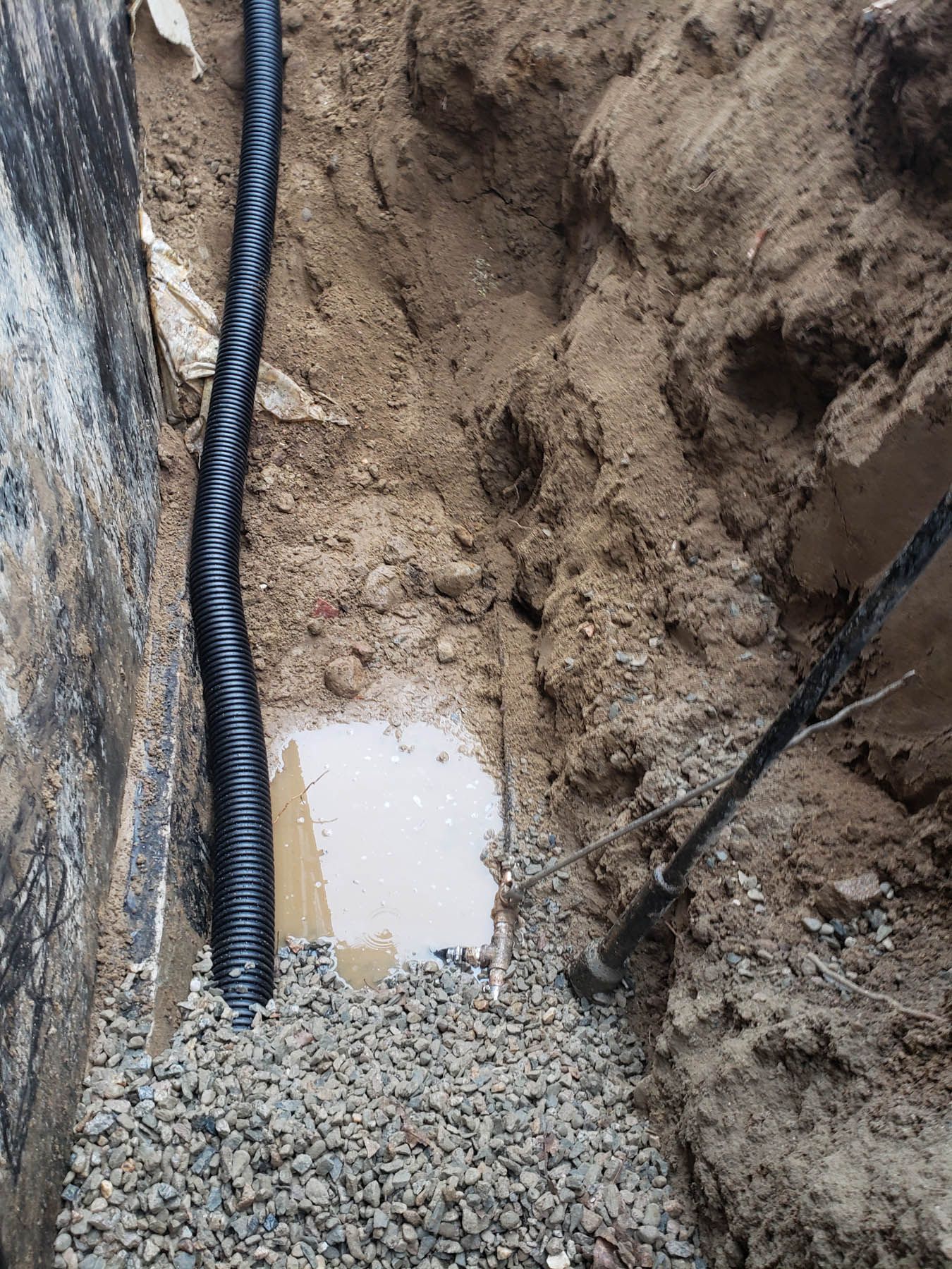 A trench next to a foundation wall, with a perforated pipe, gravel, and water.