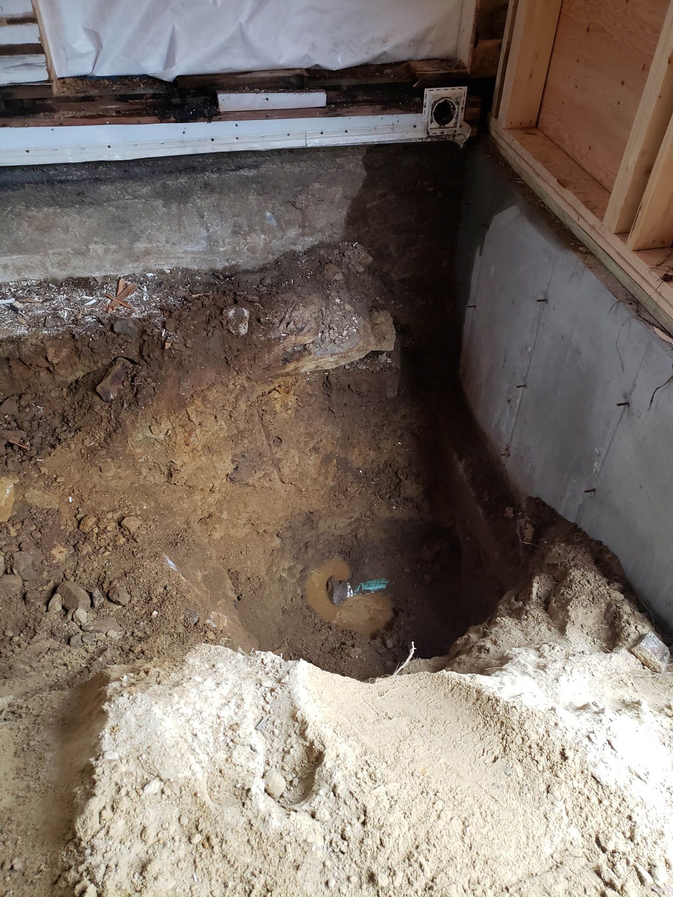Excavated area in a basement with exposed soil, pipes, and concrete foundation. Wooden framing visible.