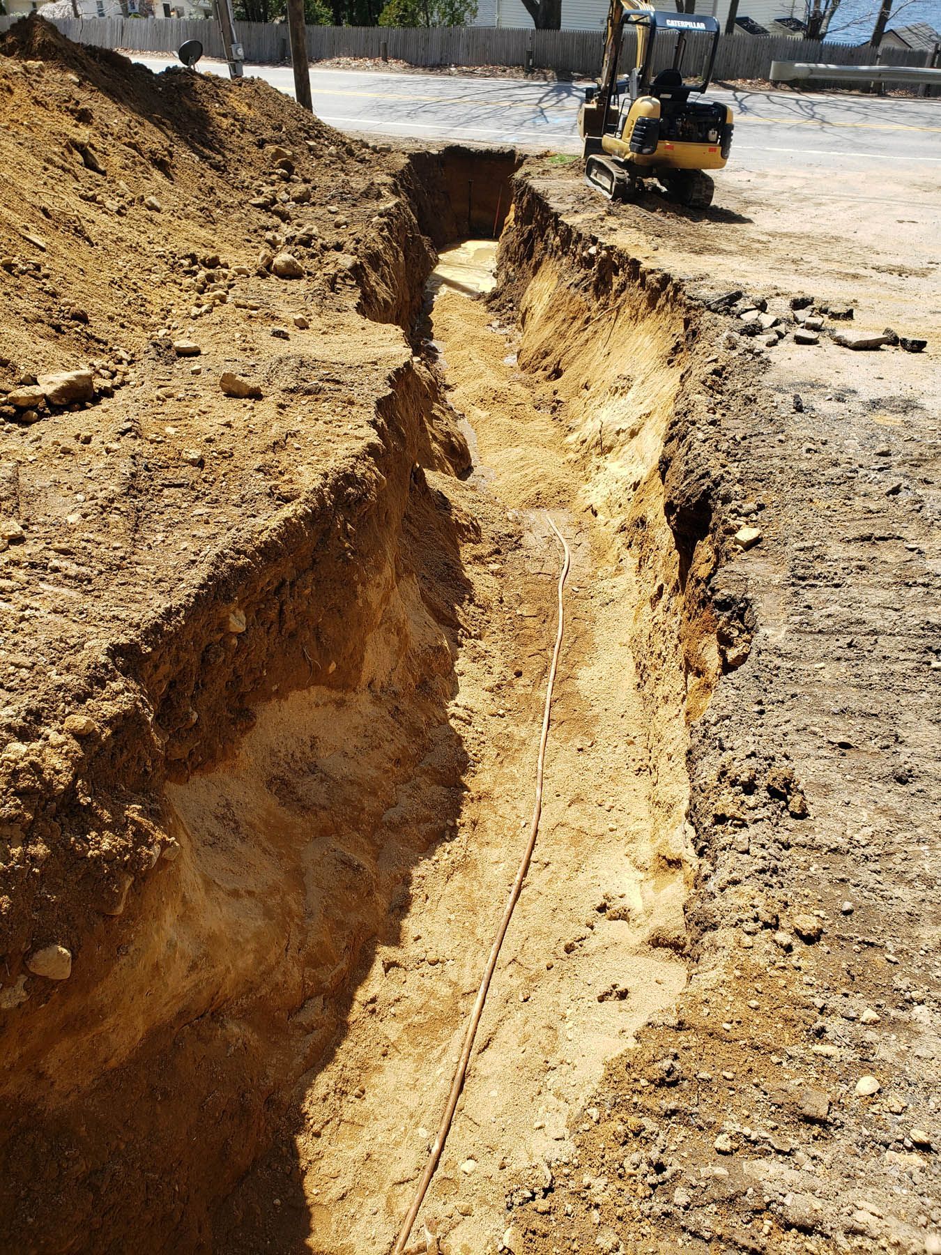 A long trench dug in the ground with a small excavator in the background, sunny day.