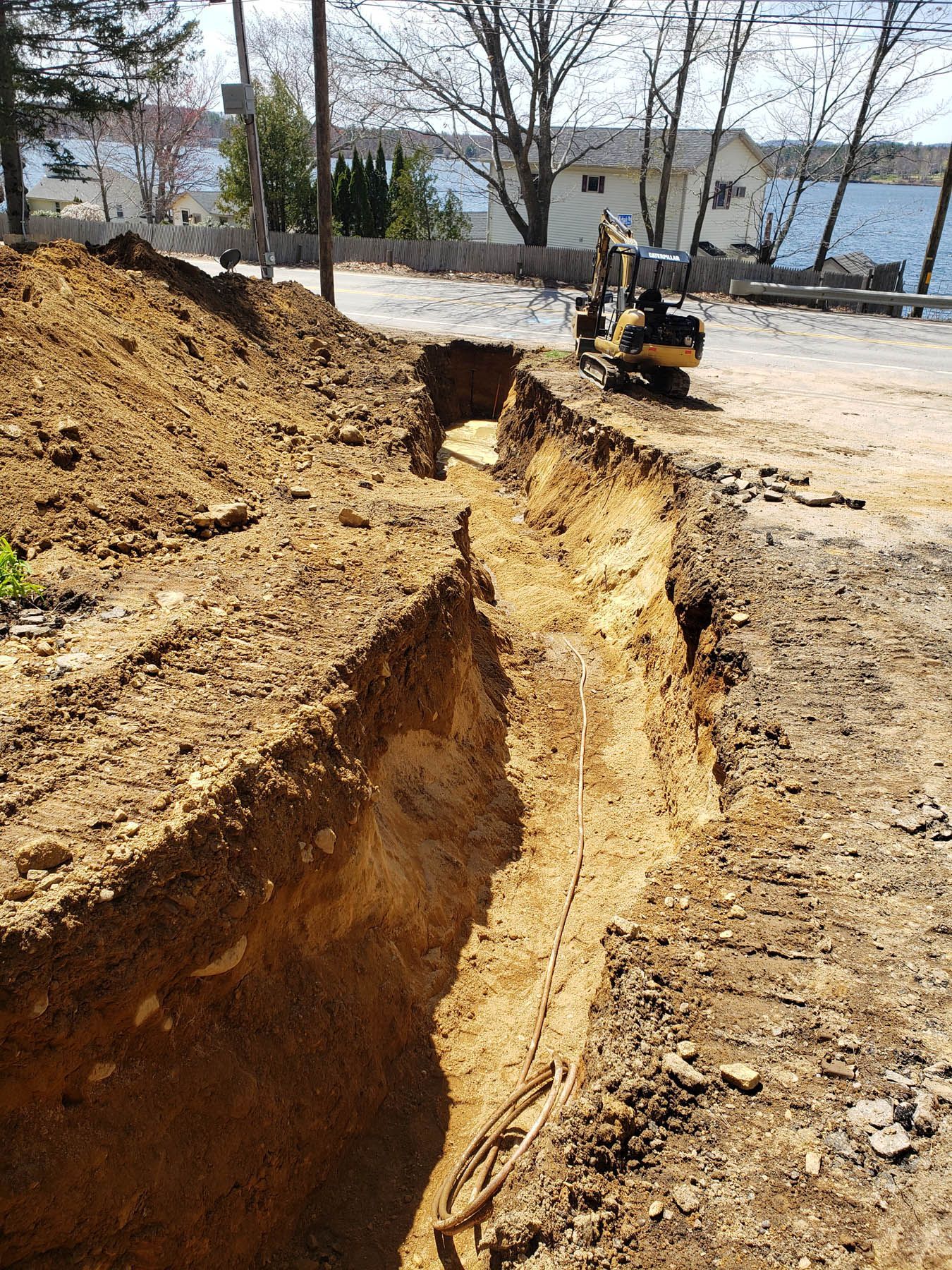 Trench dug in dirt, small excavator in background, possibly road construction.