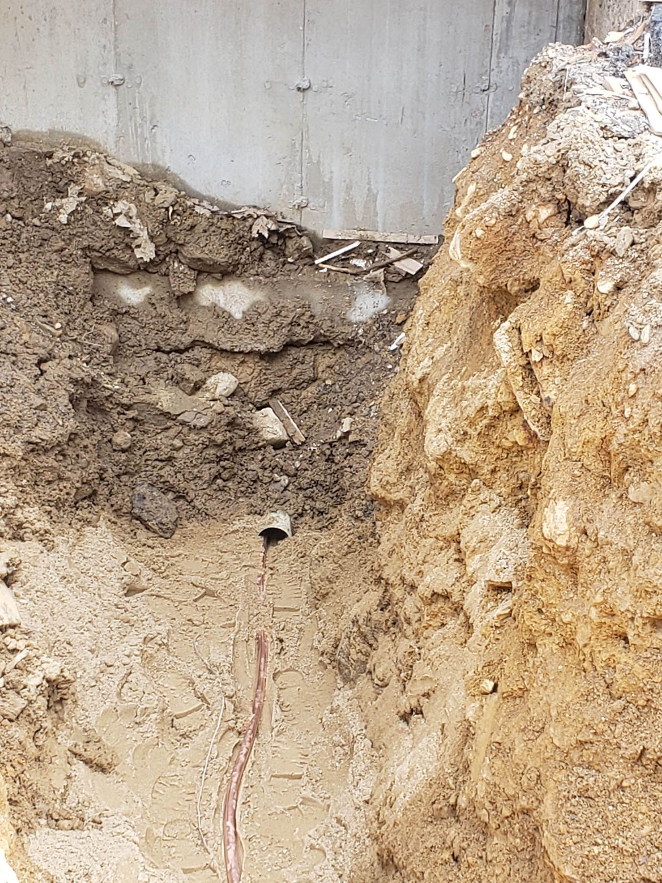Excavation pit next to a concrete wall, showing soil layers and possible utility pipe.