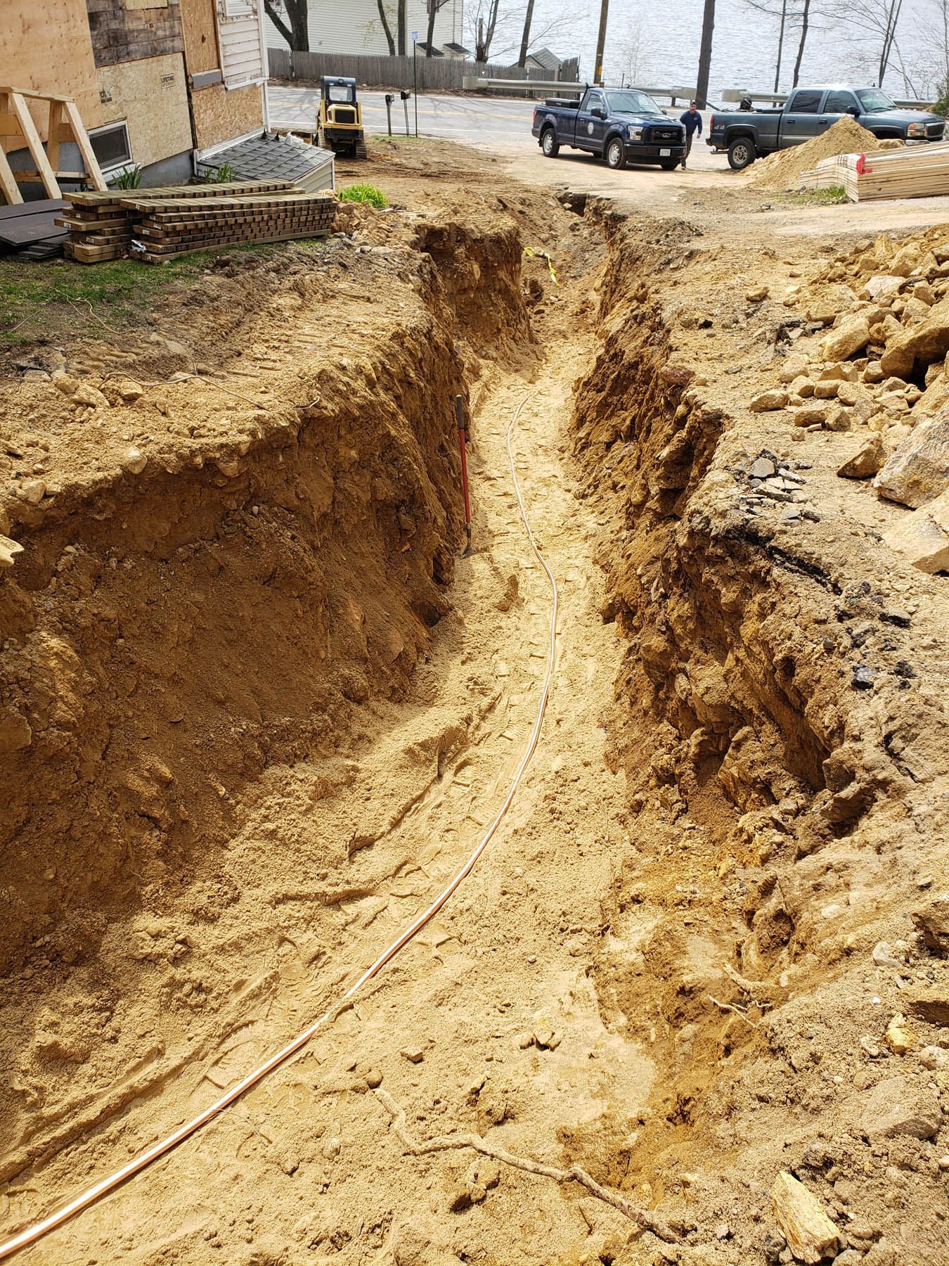 Trench dug in dirt at a construction site; two trucks, a backhoe, and a partially built house in the background.