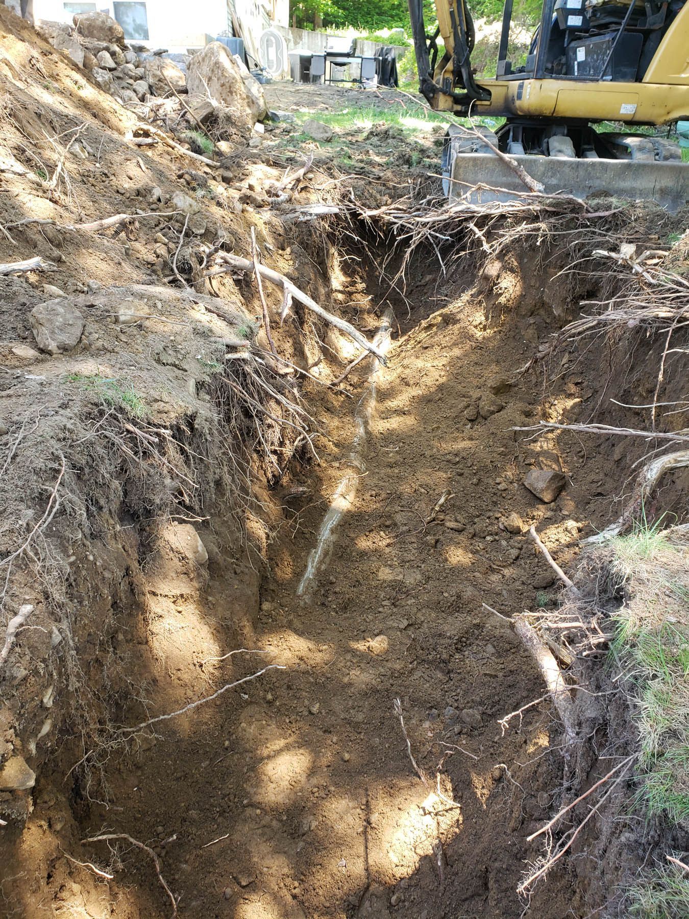 Trench dug in dirt with exposed white pipe, small excavator in the background.