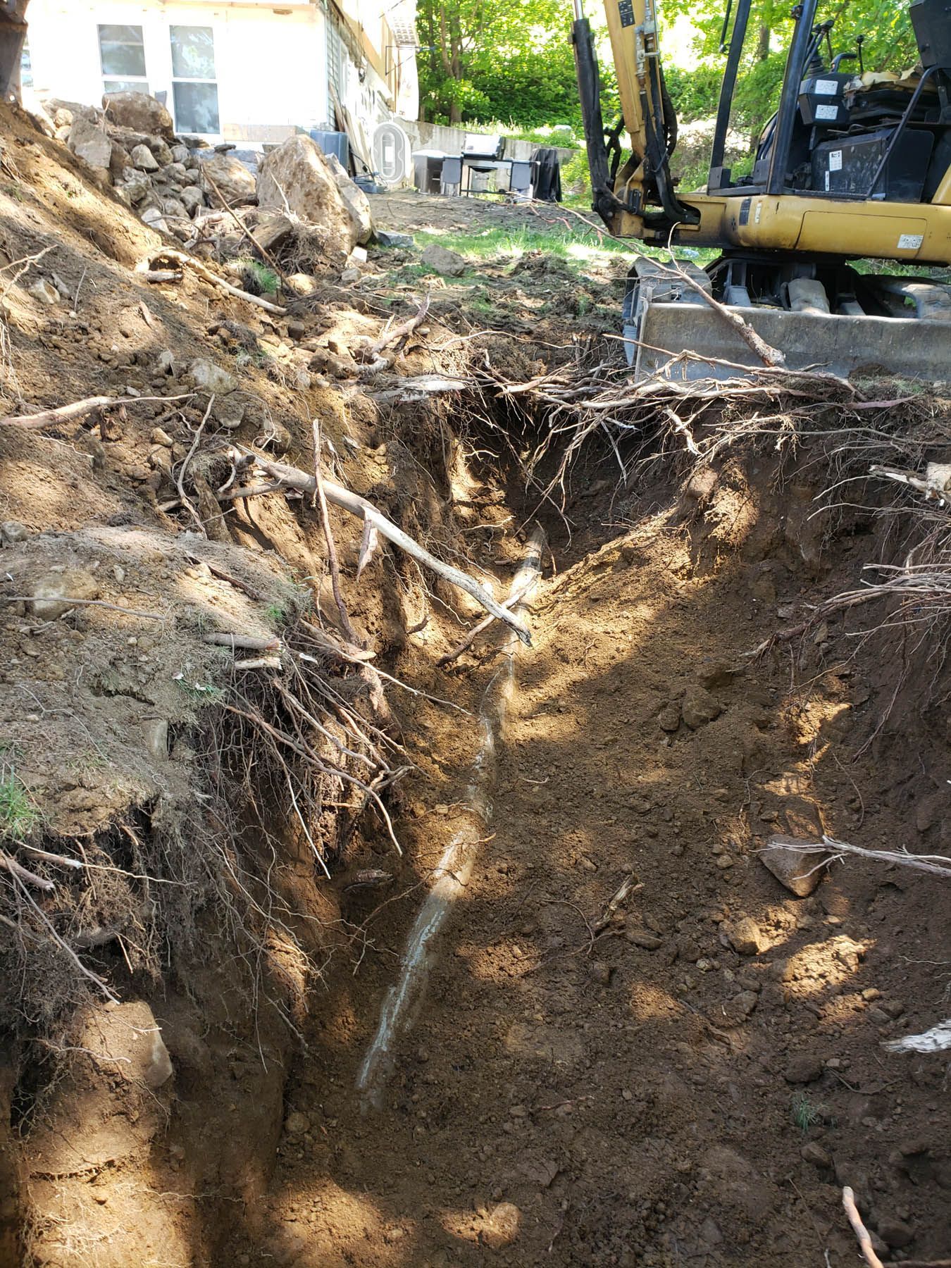 Trench dug in dirt with roots and excavator in the background, next to a house.