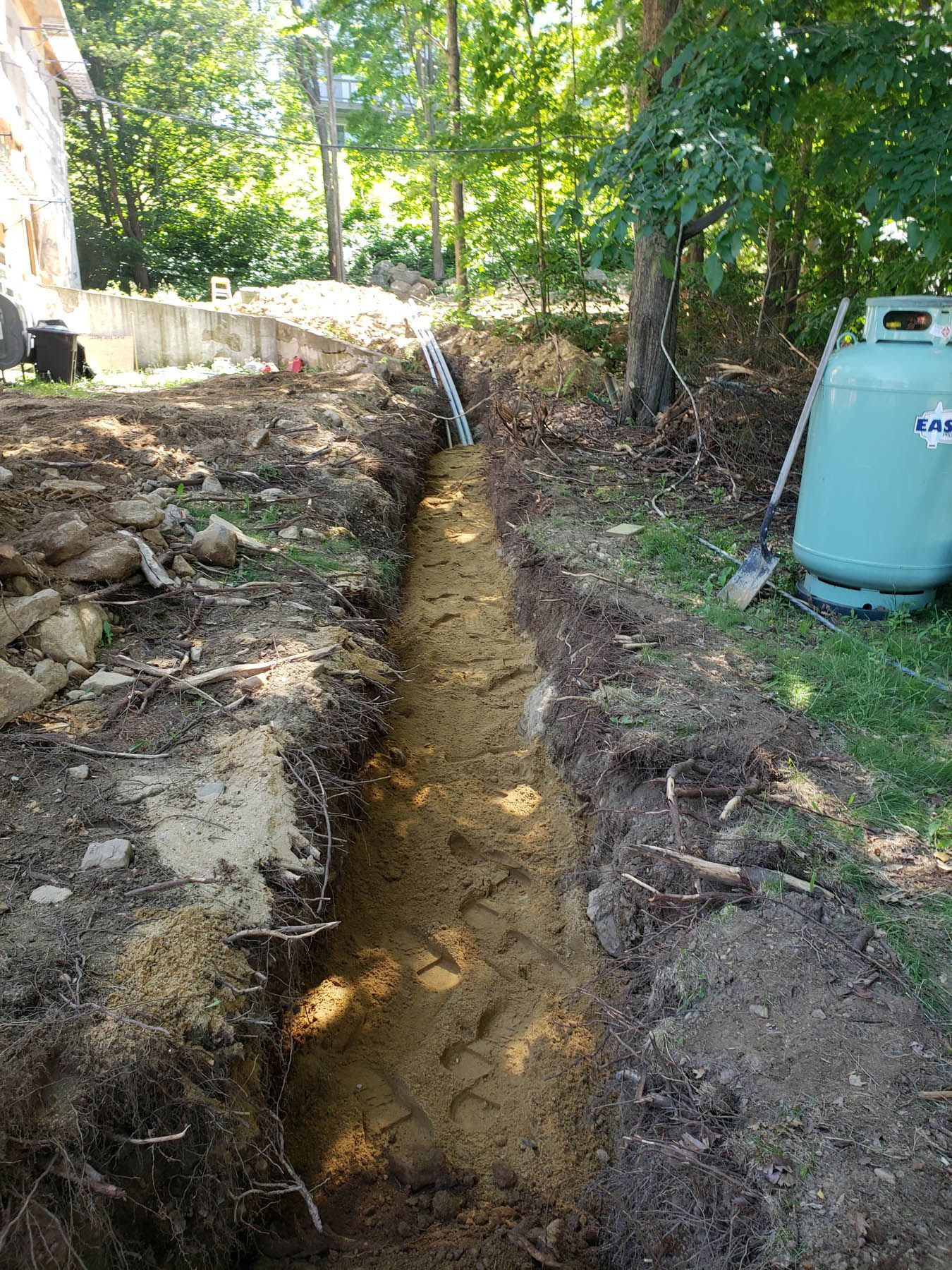 A trench dug in a yard, leading towards trees. A propane tank is in the grass nearby.