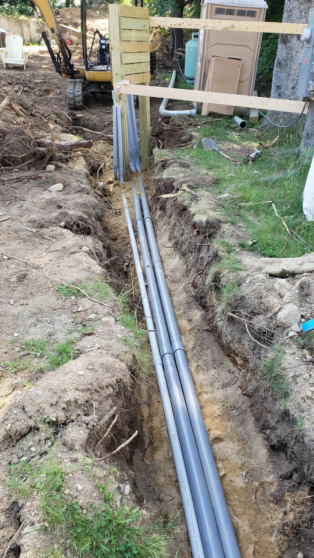 Trench with gray pipes running alongside a wooden structure, likely for underground utilities.