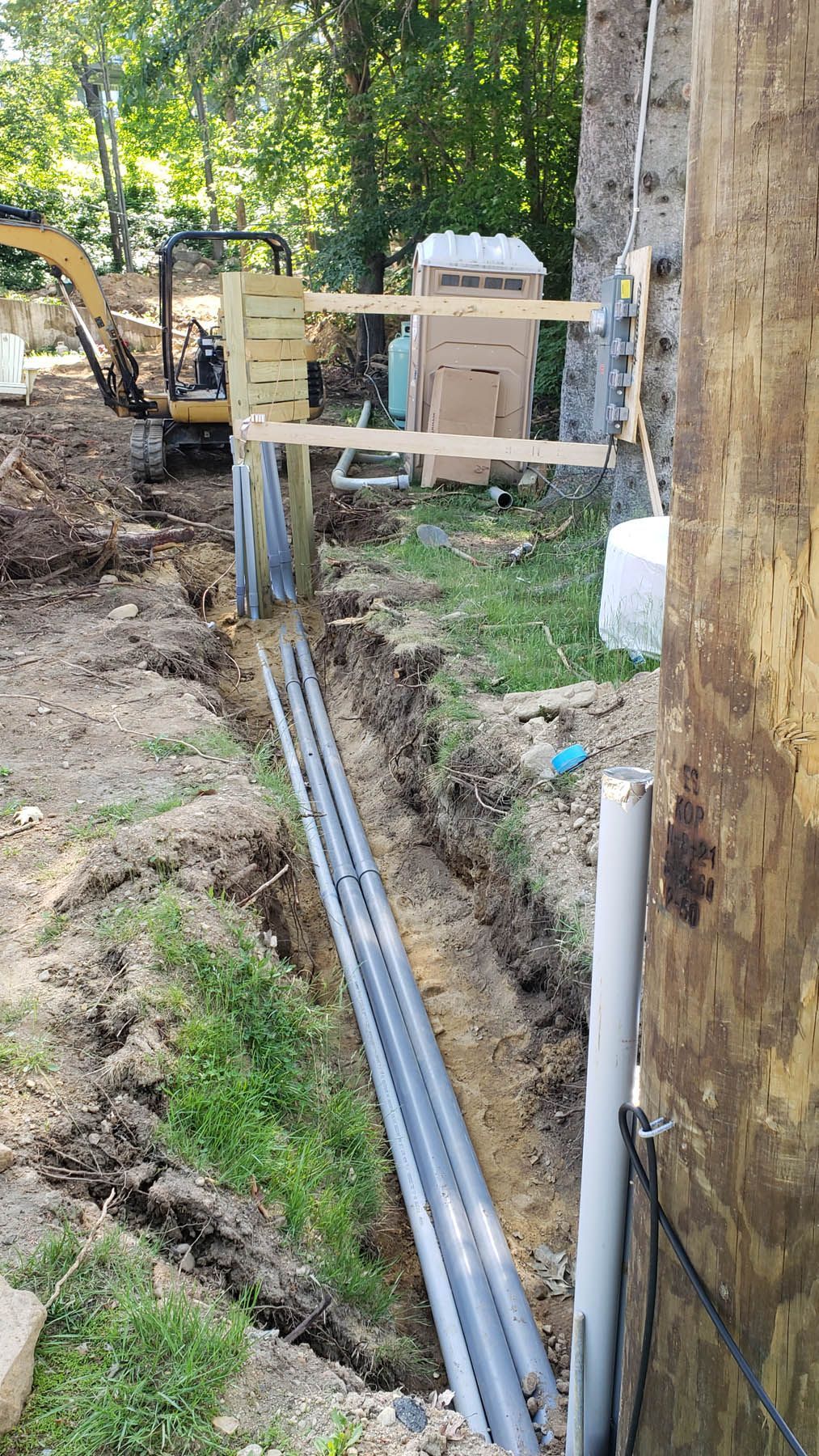 Trench with gray pipes, next to a wall and mini excavator, trees in the background.