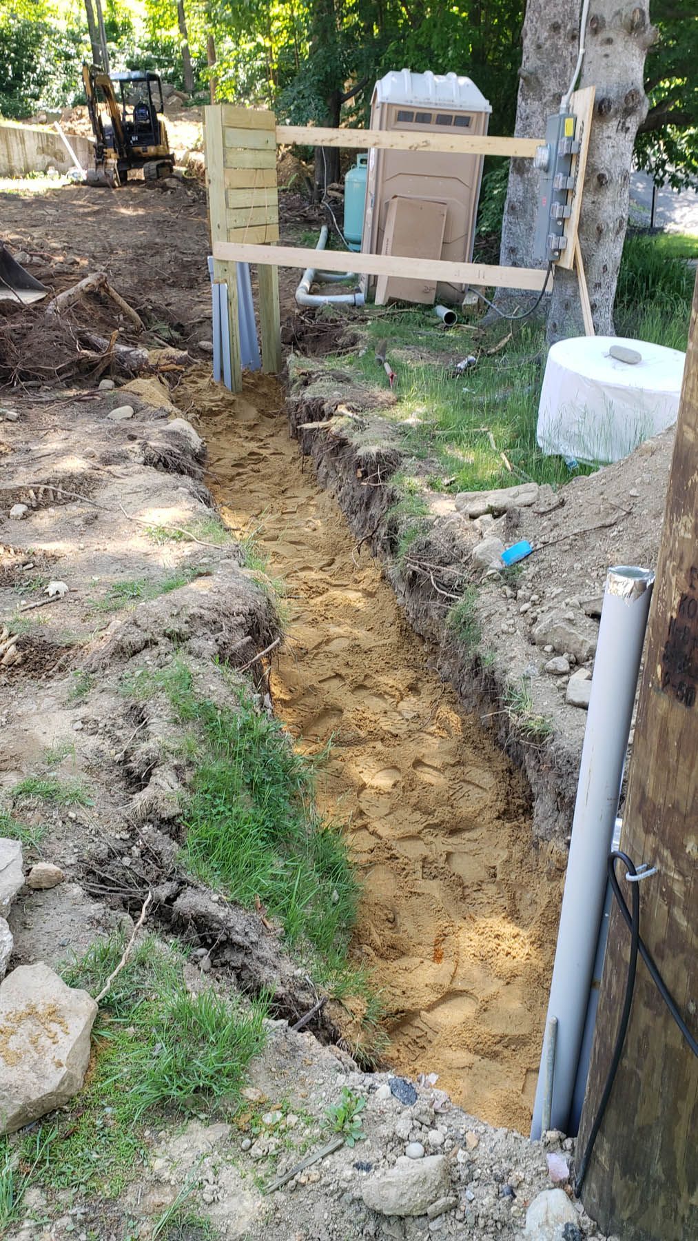A long trench dug for construction, with wood supports, a portable toilet, and a small bulldozer in the background.