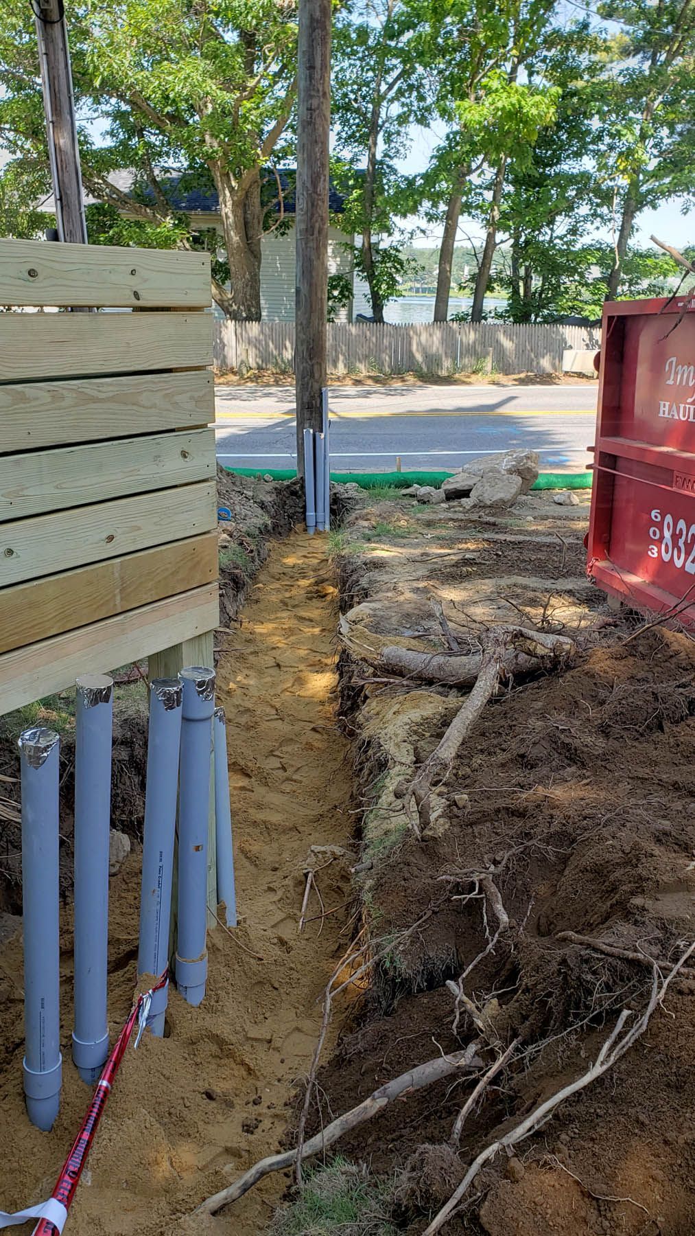 Trench dug near a wooden fence. Gray PVC pipes are in the trench, with dirt and roots visible.
