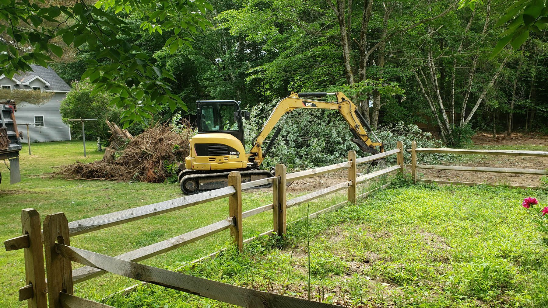 Yellow excavator clearing brush behind a wooden fence.