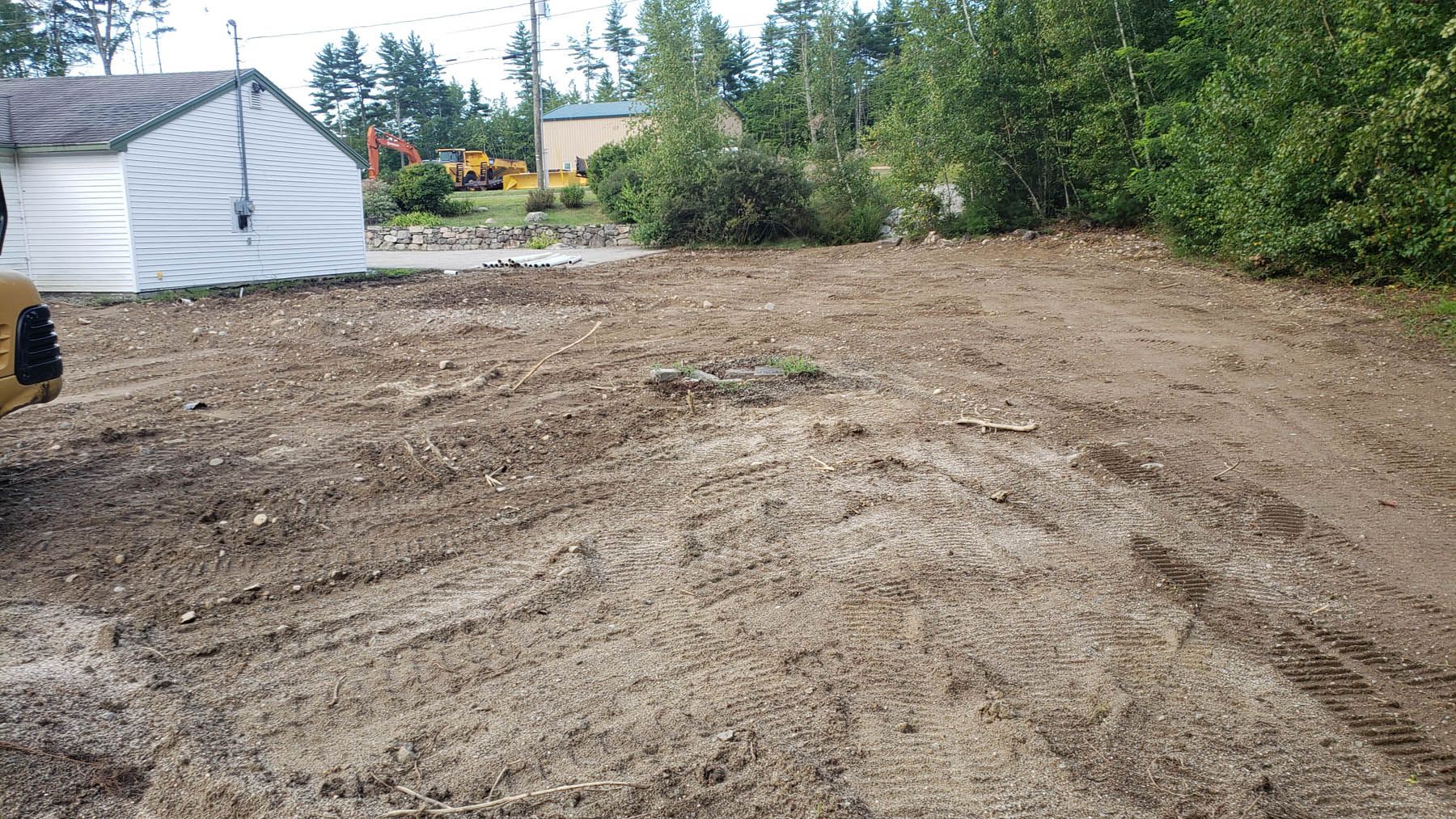 A dirt lot with tire tracks, a small white building on the left, and trees in the background.