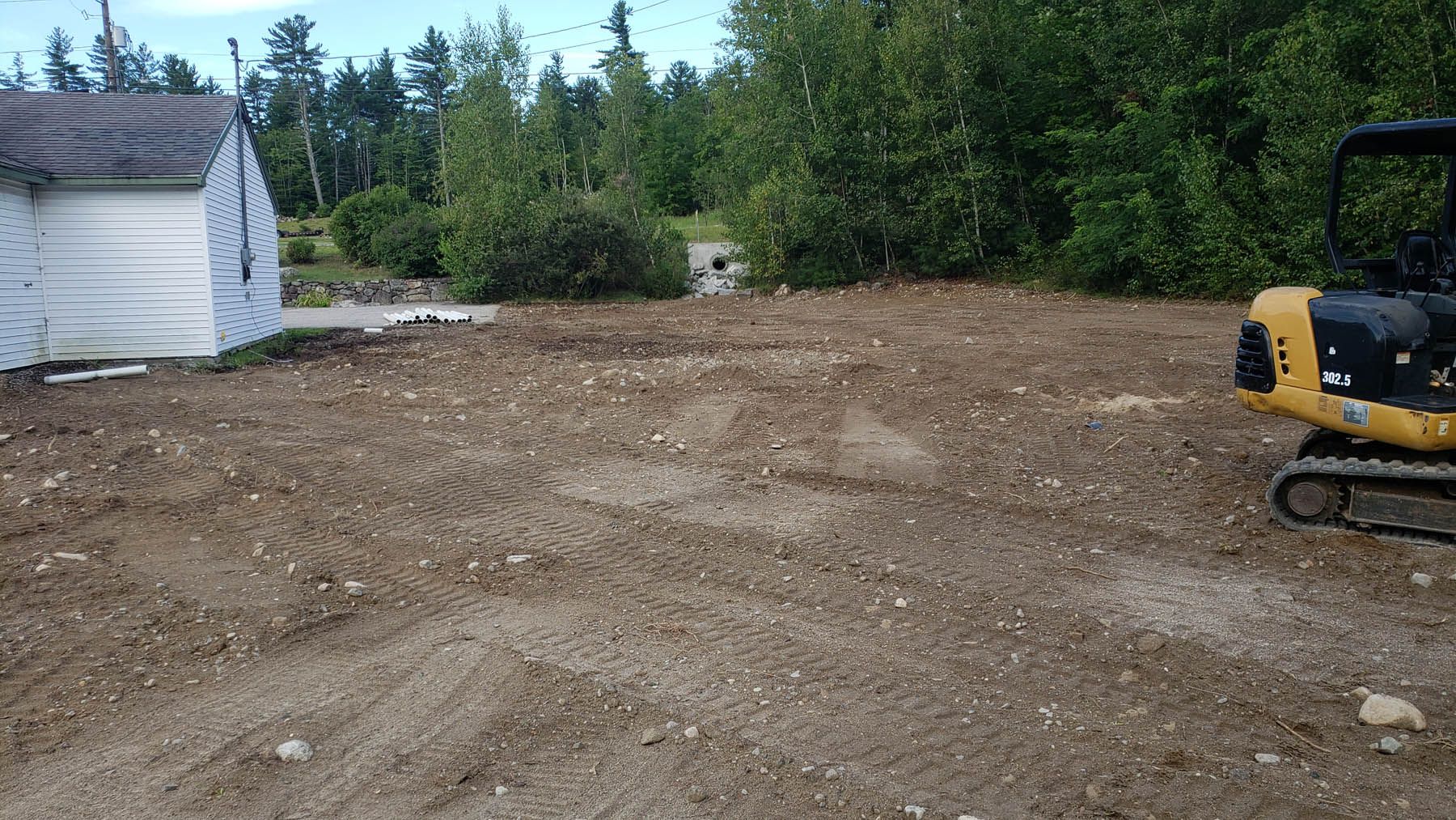 Dirt lot with an excavator next to a small building and treeline.