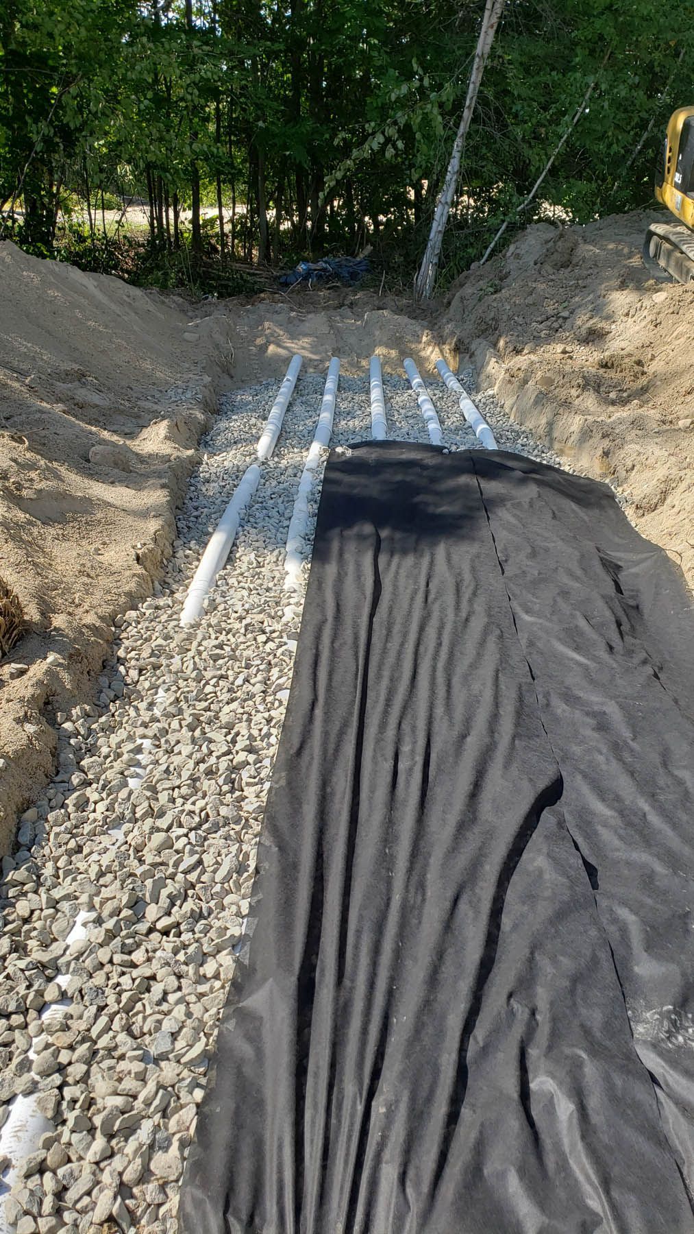 Trench with gravel, white pipes, and black fabric, likely for drainage or septic system construction, outdoors.