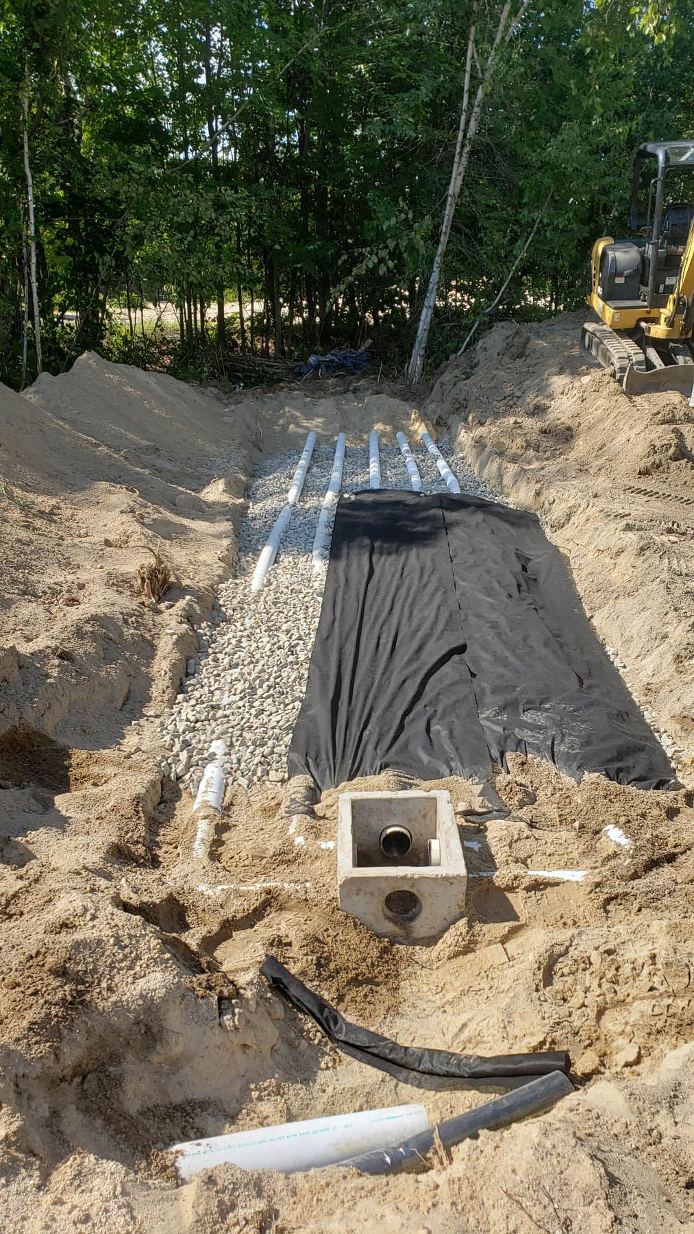 Septic system installation: trench with gravel, fabric, pipes, and concrete distribution box. Sunlight illuminates the scene.