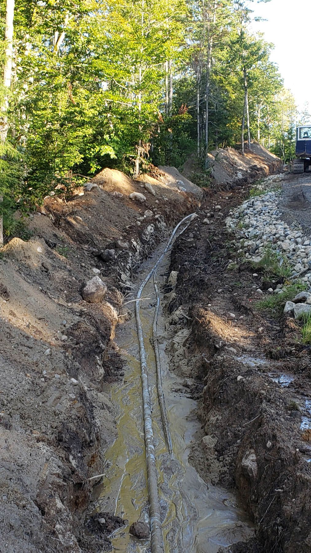 Trench dug in muddy dirt with buried pipes, beside a gravel road, trees in background.