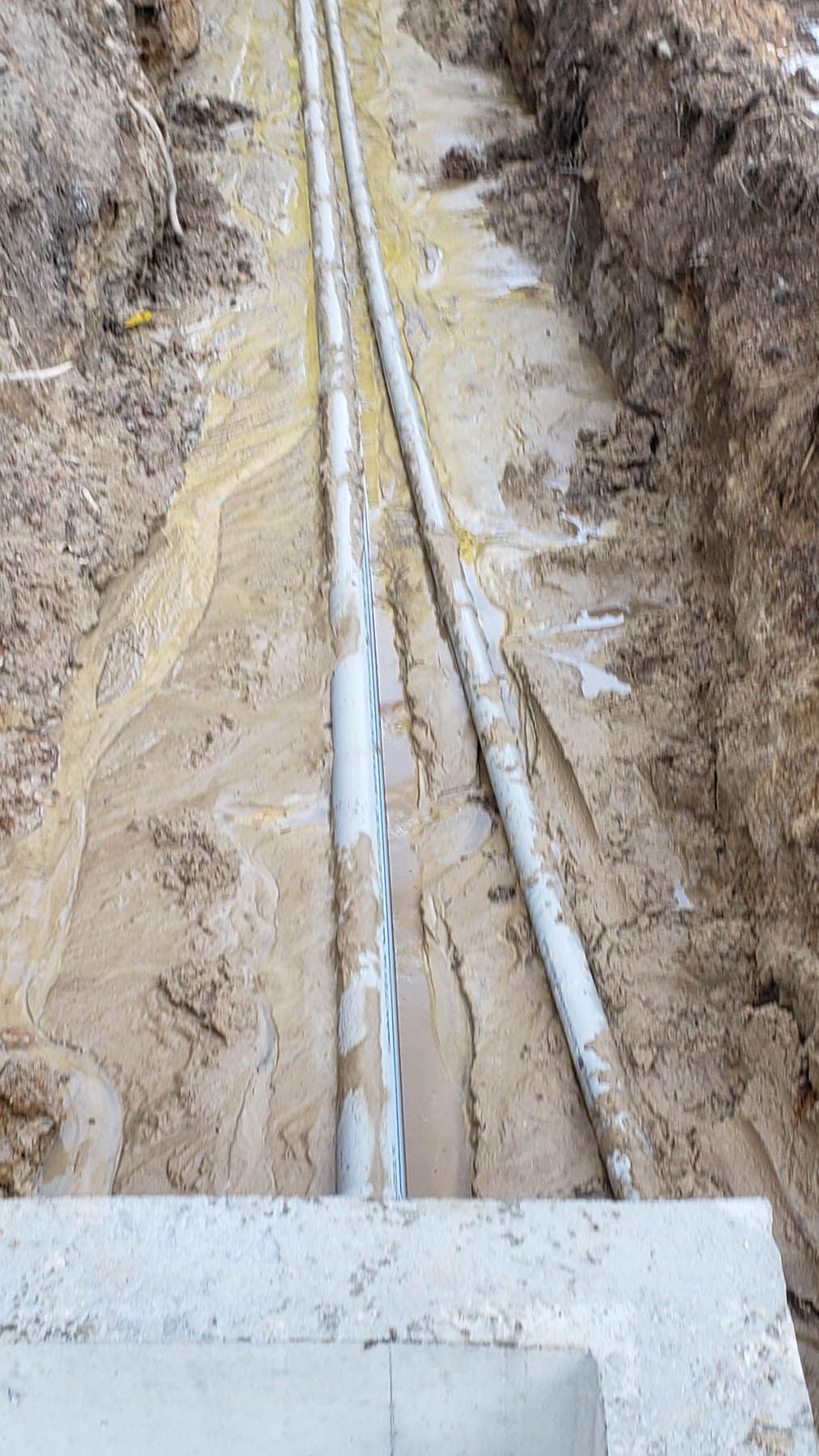 Trench with two grey pipes laid in wet dirt, a concrete structure is in the foreground.