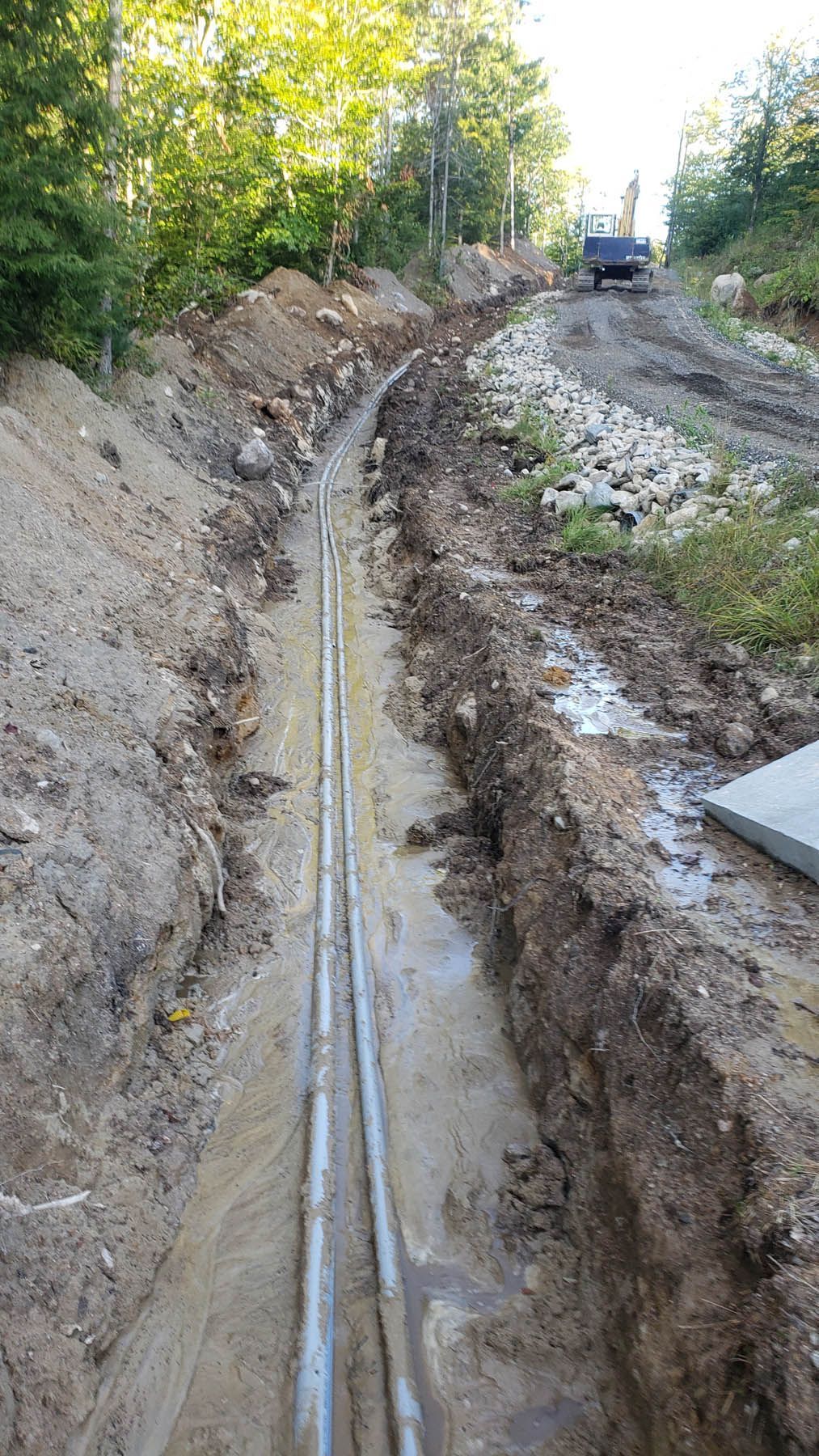 Trench with metal pipes, running along a dirt road in a wooded area. A truck is at the top of the road.