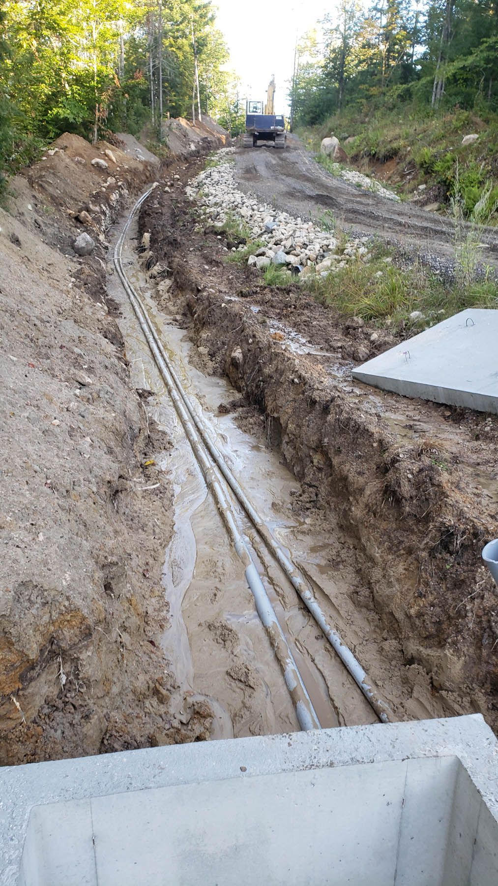 Construction site: Trench with pipes, earth, and a truck on a dirt road surrounded by trees.