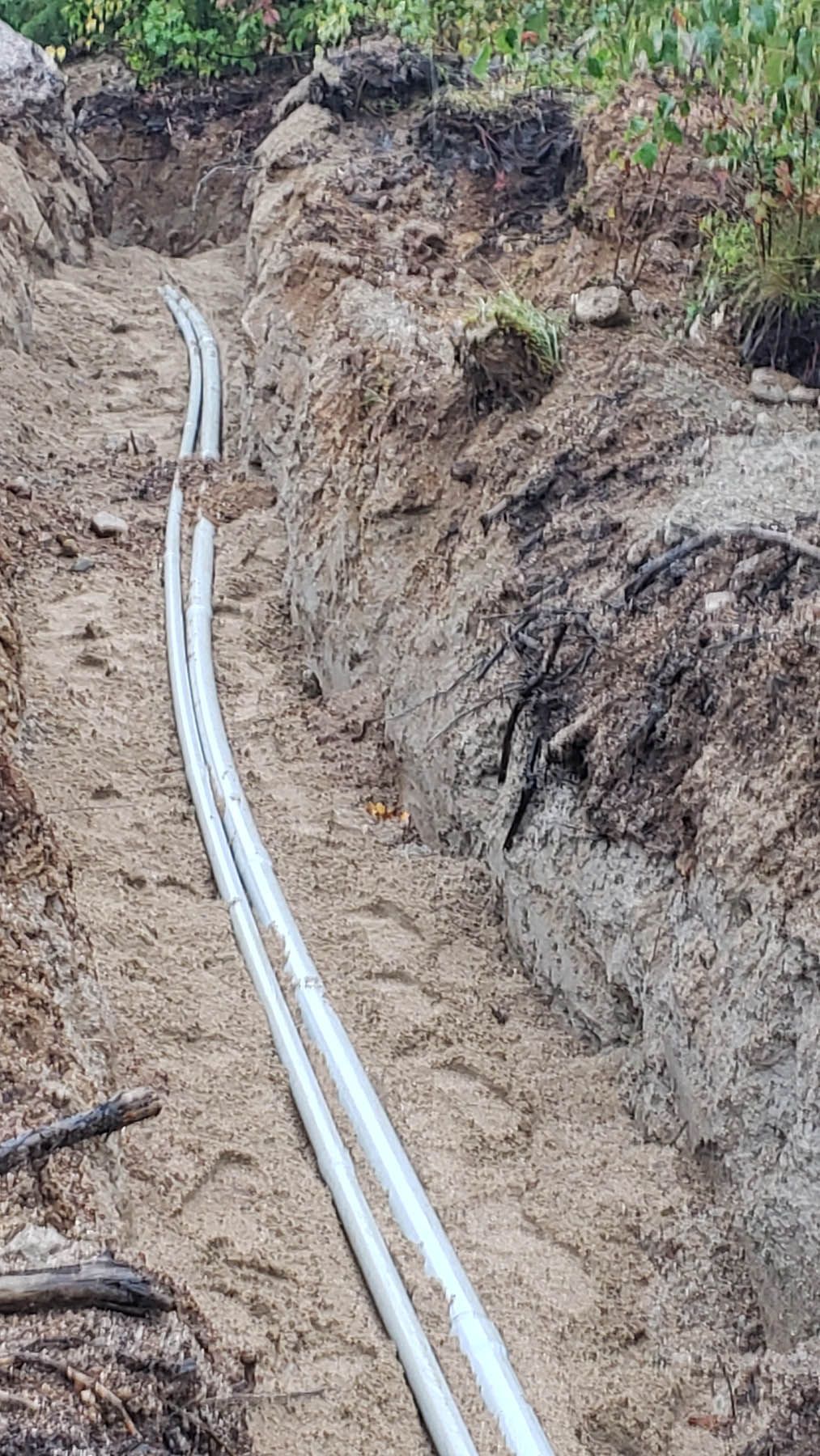 Two white pipes laid in a narrow, sandy trench; dirt walls in an outdoor setting.