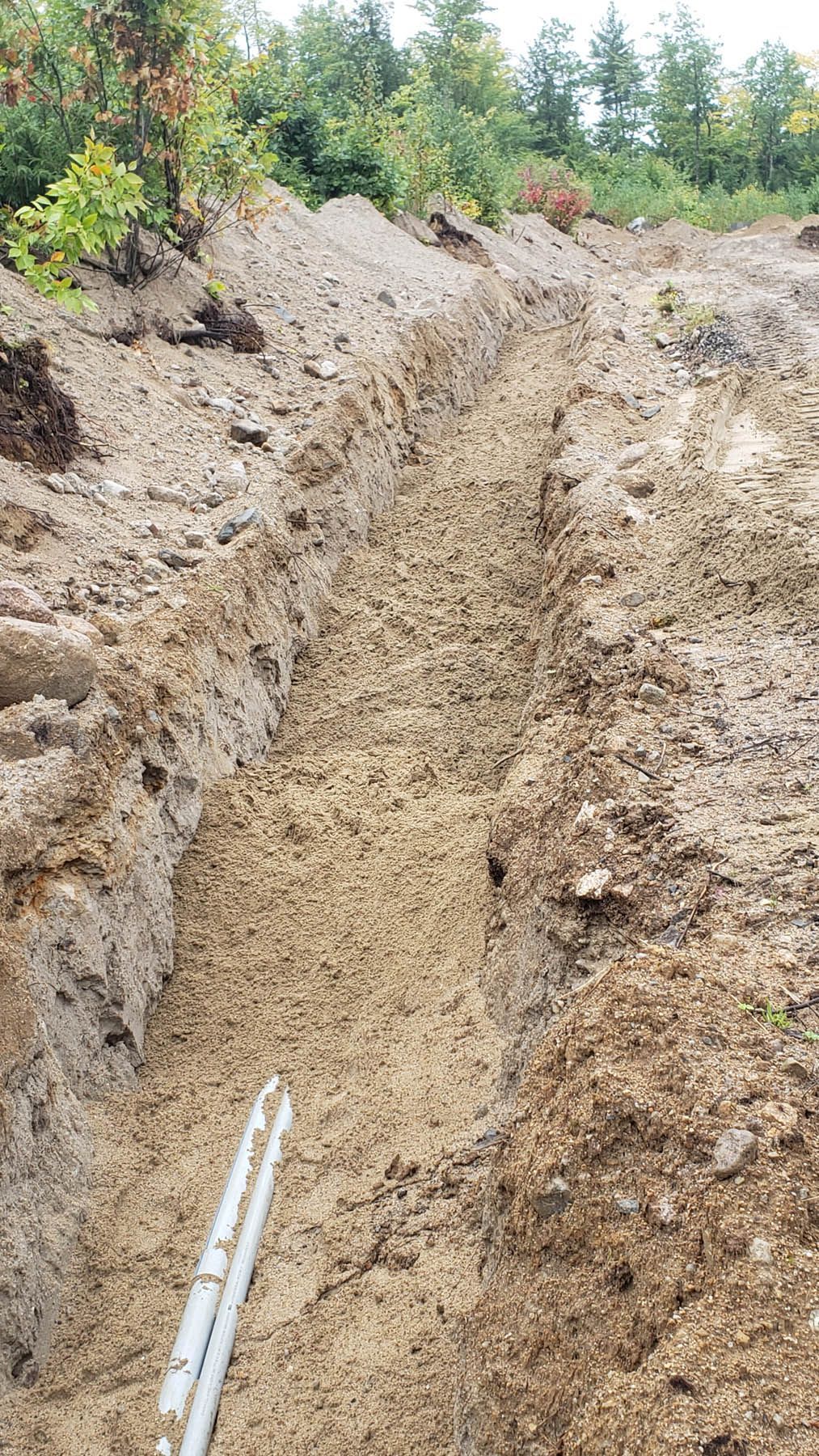 Long, narrow trench dug in dirt, two white pipes visible. Dirt and trees in the background.