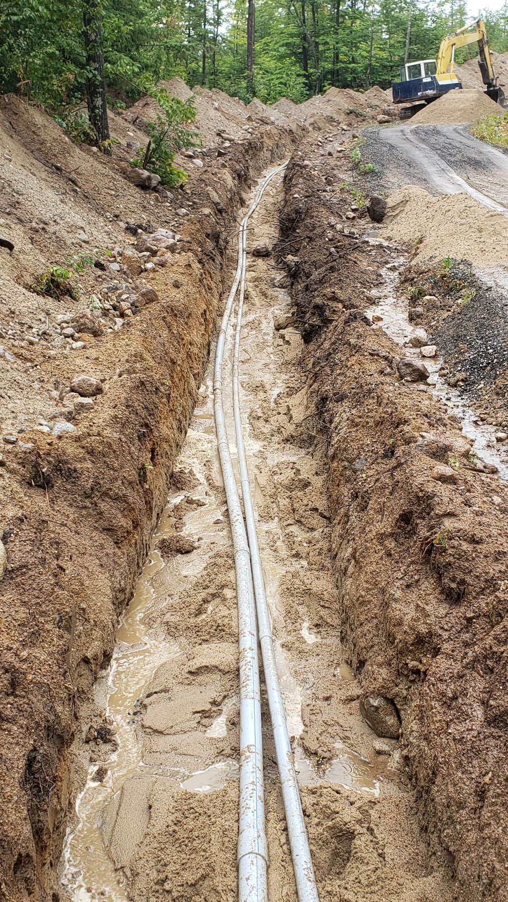 A long, muddy trench with parallel gray pipes, likely for utility installation, near a construction site.