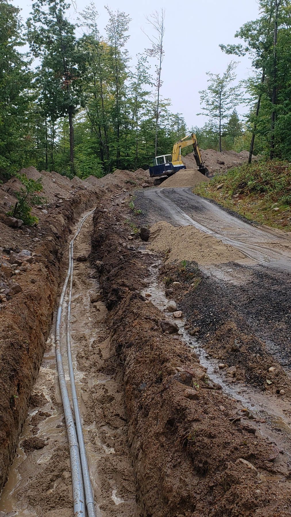 Trench dug in dirt with buried cables. Excavator works in the background, surrounded by trees.