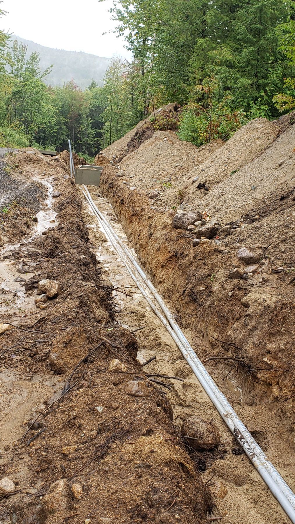 Trench with white pipes running through it. Dirt piles border the sides, trees in the background.