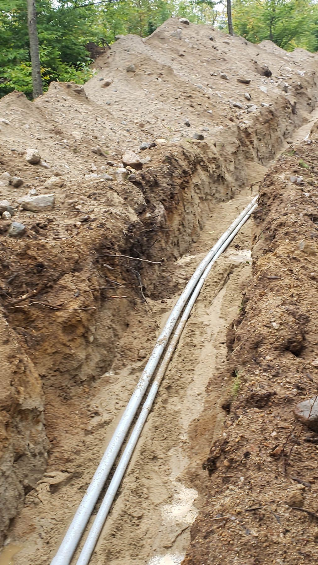 Trench dug in dirt with two gray pipes laid inside, likely for utilities, with a large pile of dirt beside.