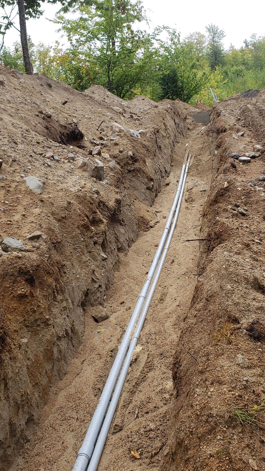 Two gray pipes laid in a narrow, freshly dug trench in a dirt hillside; trees in the background.