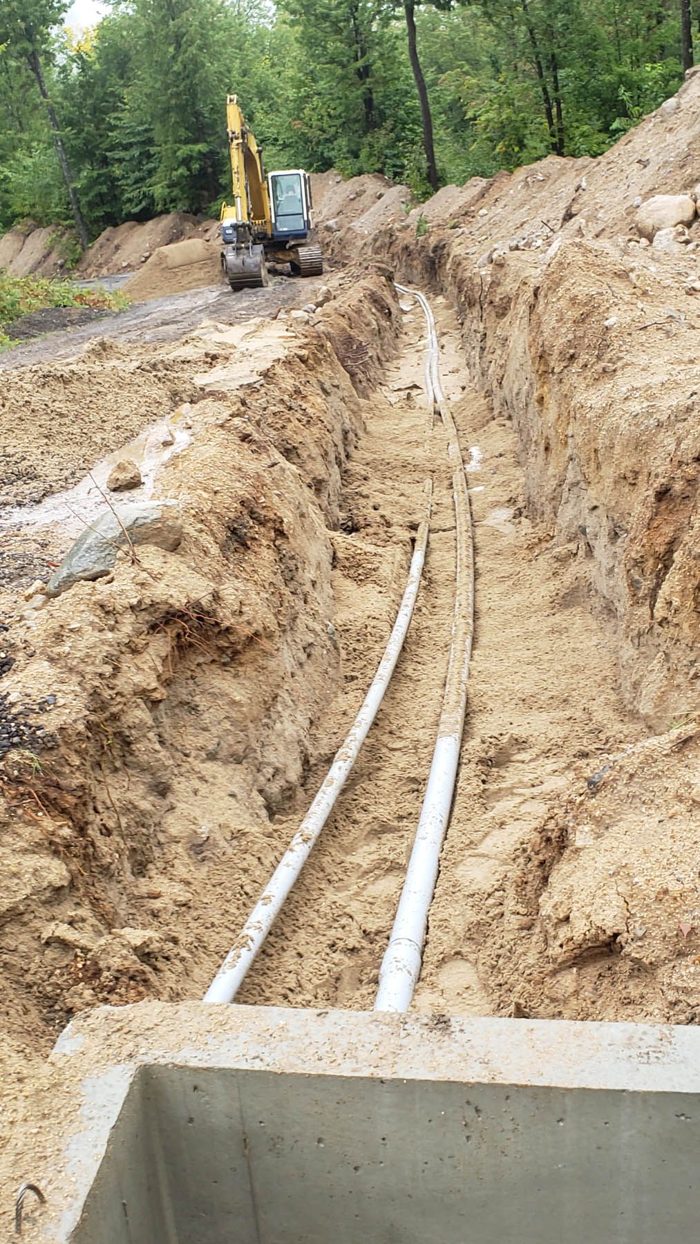 Two white pipes in a trench with an excavator in the background, construction site.