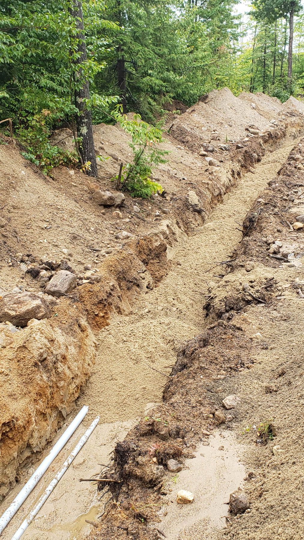 A long, narrow trench dug in dirt, filled with gravel, with pipes visible. Green trees in the background.