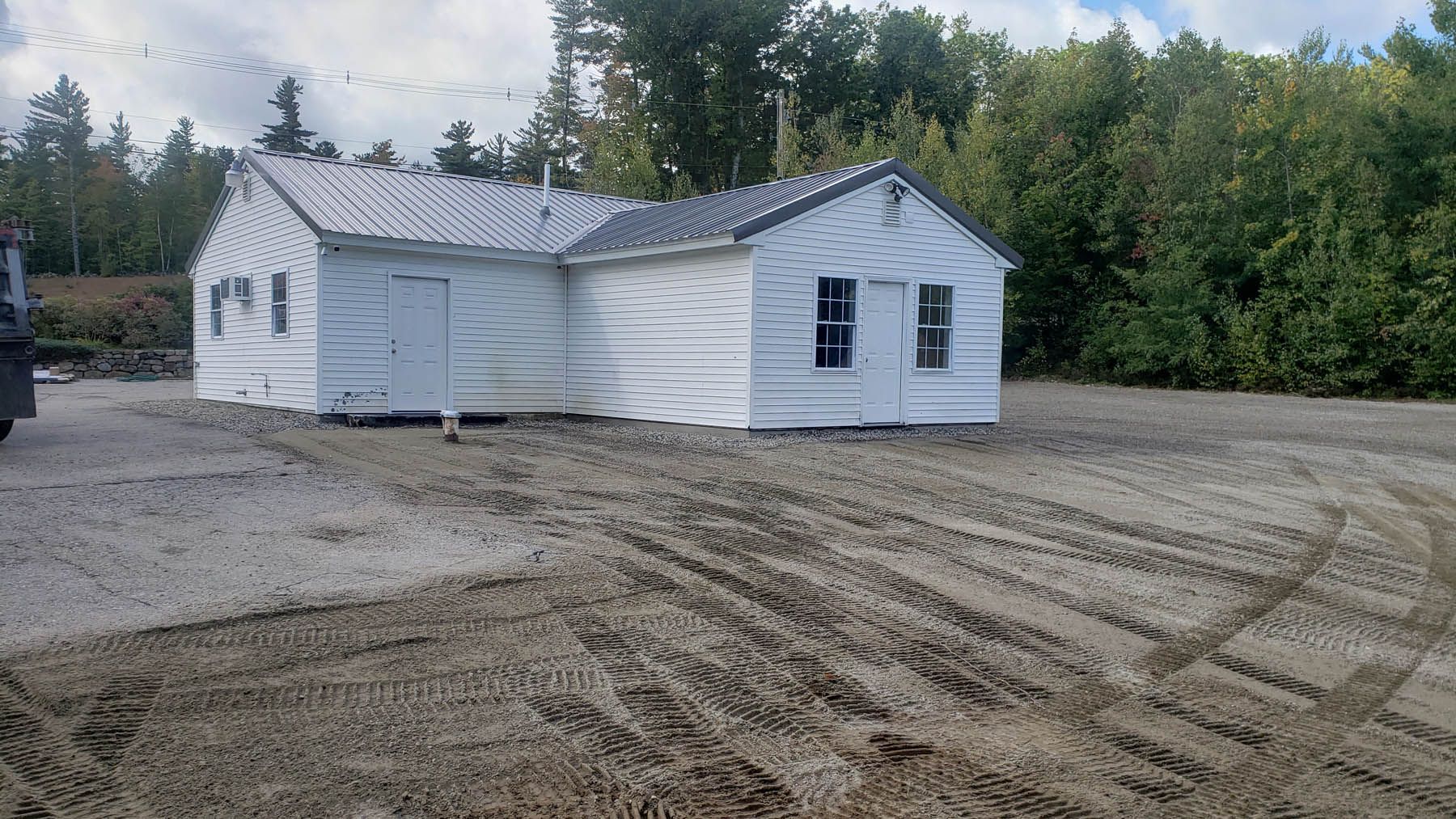 White-painted small house on a gravel lot, with trees in the background. Tire tracks in the foreground.