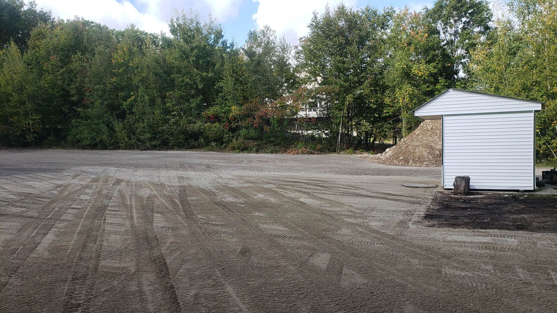 A gravel lot with a small white shed, pile of rocks, and trees in the background on a sunny day.